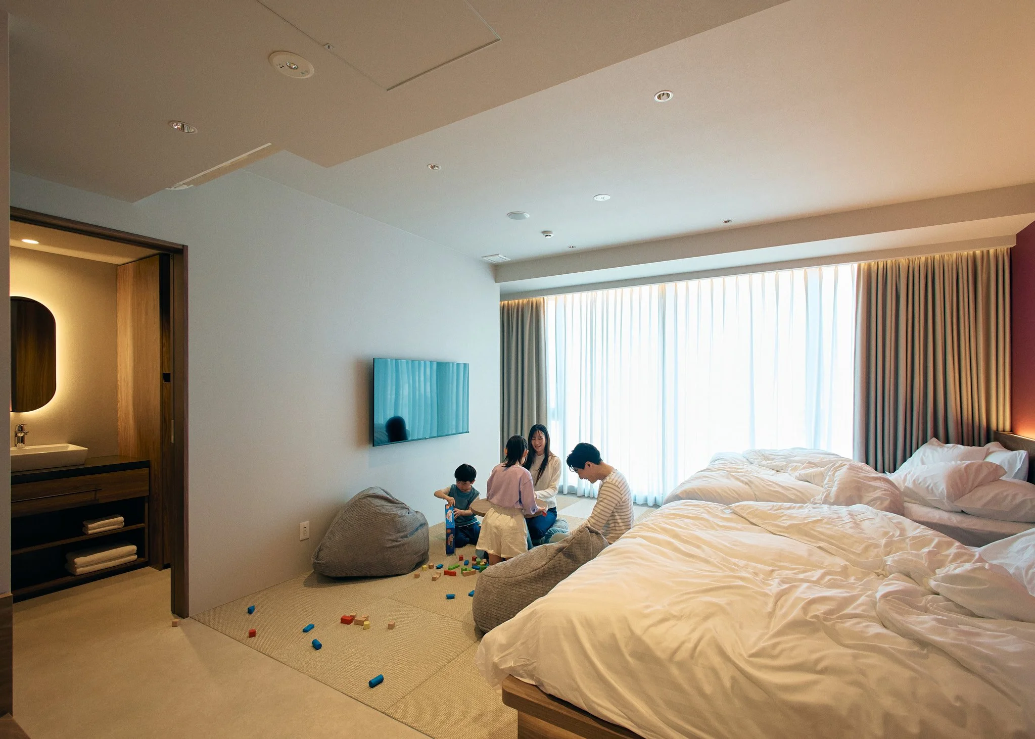 A hotel room with a large unmade bed, a flat-screen TV on the wall, and a small group of people, including children, sitting on the floor playing with toys near the window.