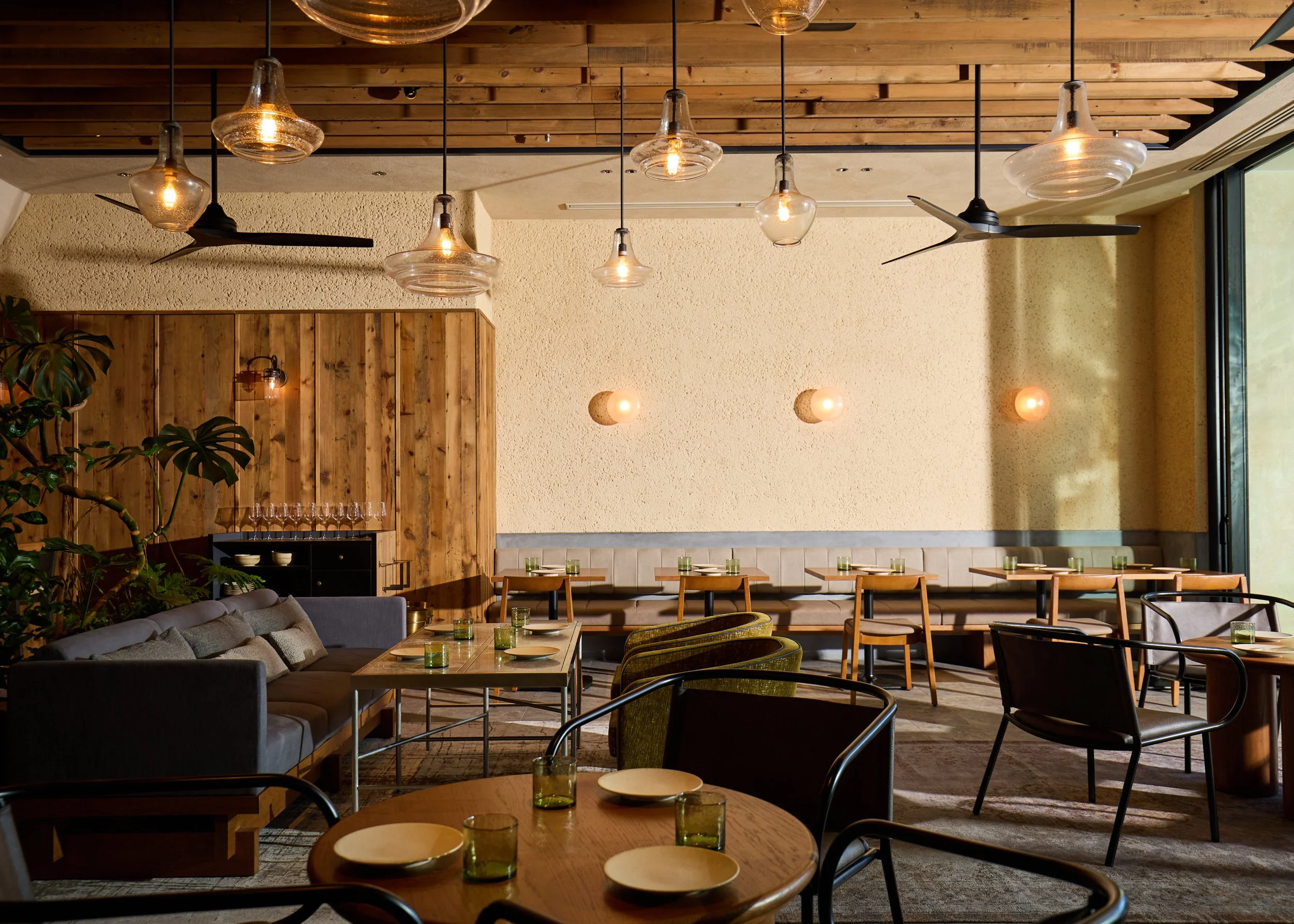 Interior of a modern restaurant with wooden ceilings, pendant lights, a mix of chairs and sofas, and a large window.