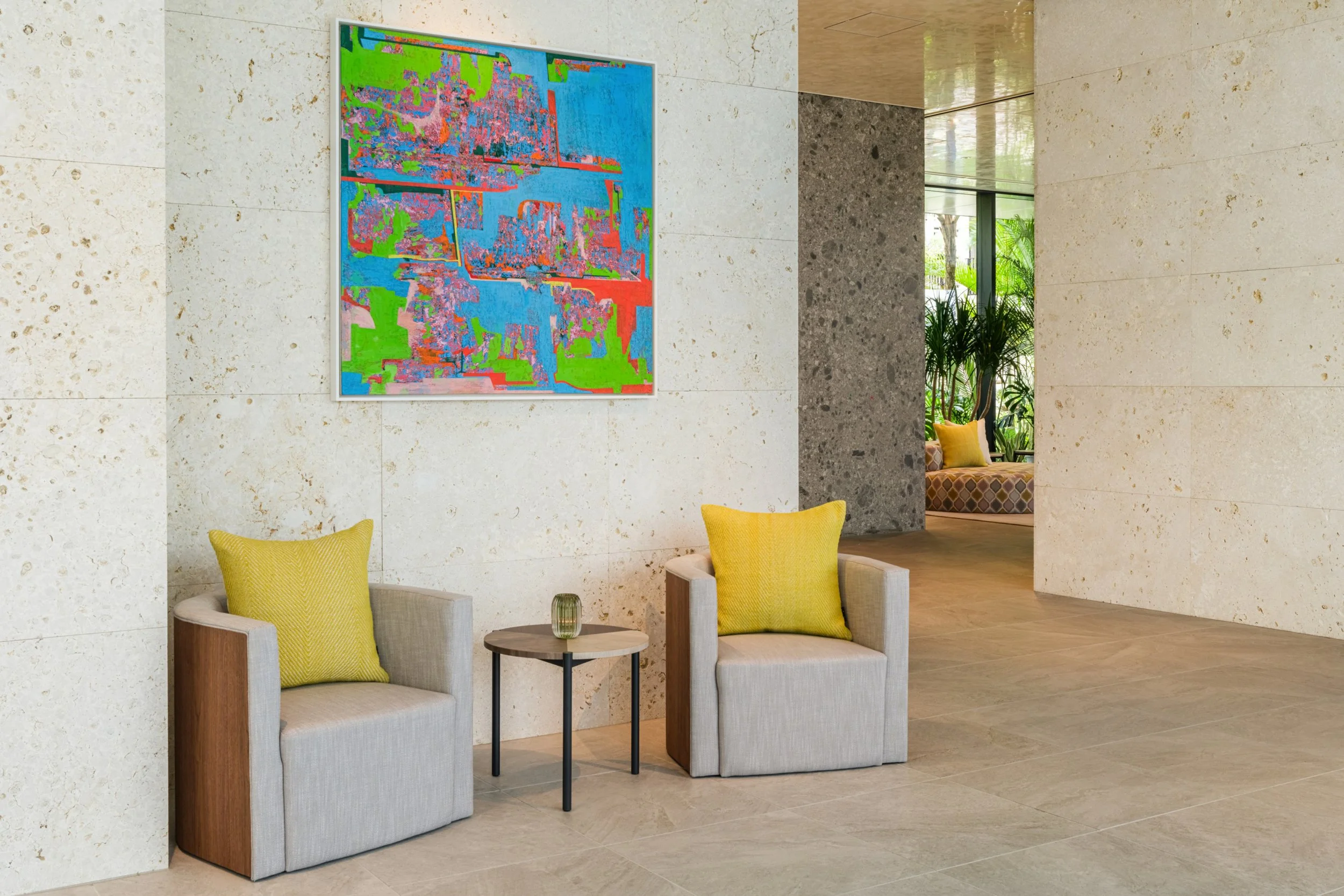 Modern lobby area with two beige armchairs with yellow pillows, a small round black side table with a green vase, white stone walls, a colorful abstract painting, and large windows with green plants visible outside.