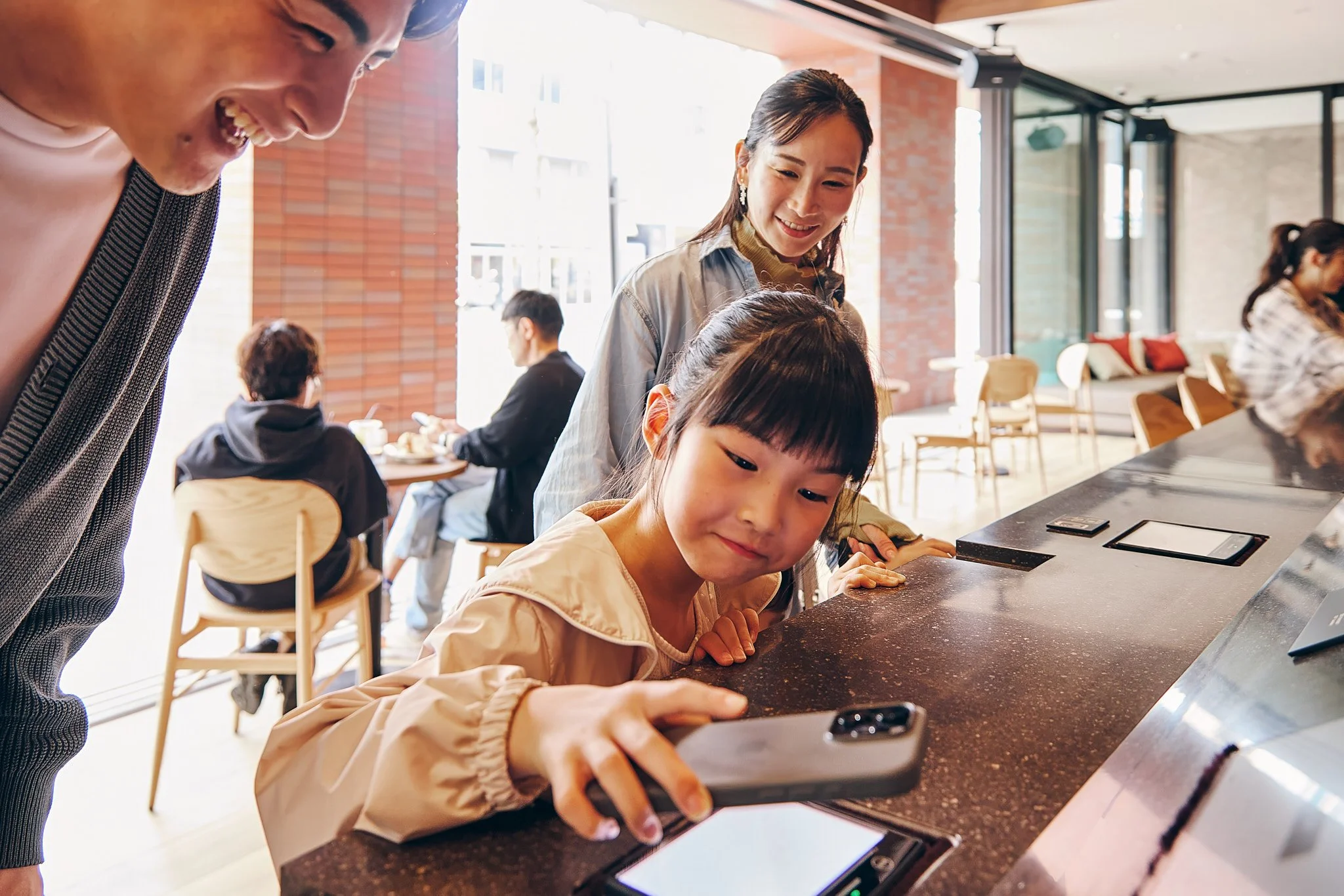 A young girl takes a selfie with a smartphone at a cafe counter, with an adult woman and a man smiling nearby. In the background, other people are sitting at tables.