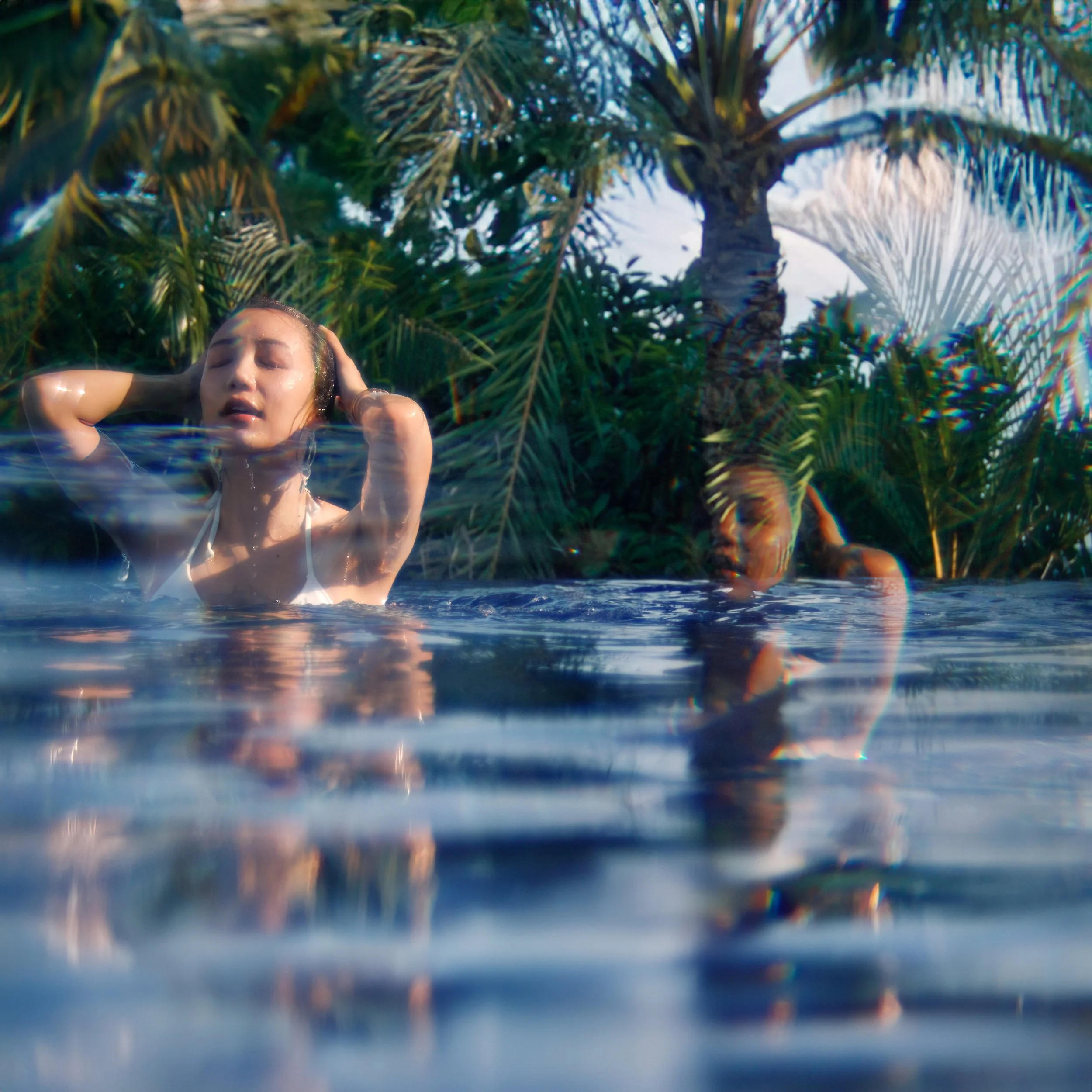 Two women relaxing in a pool surrounded by tropical palm trees, with one woman submerged up to her shoulders, eyes closed, and hands in her hair.