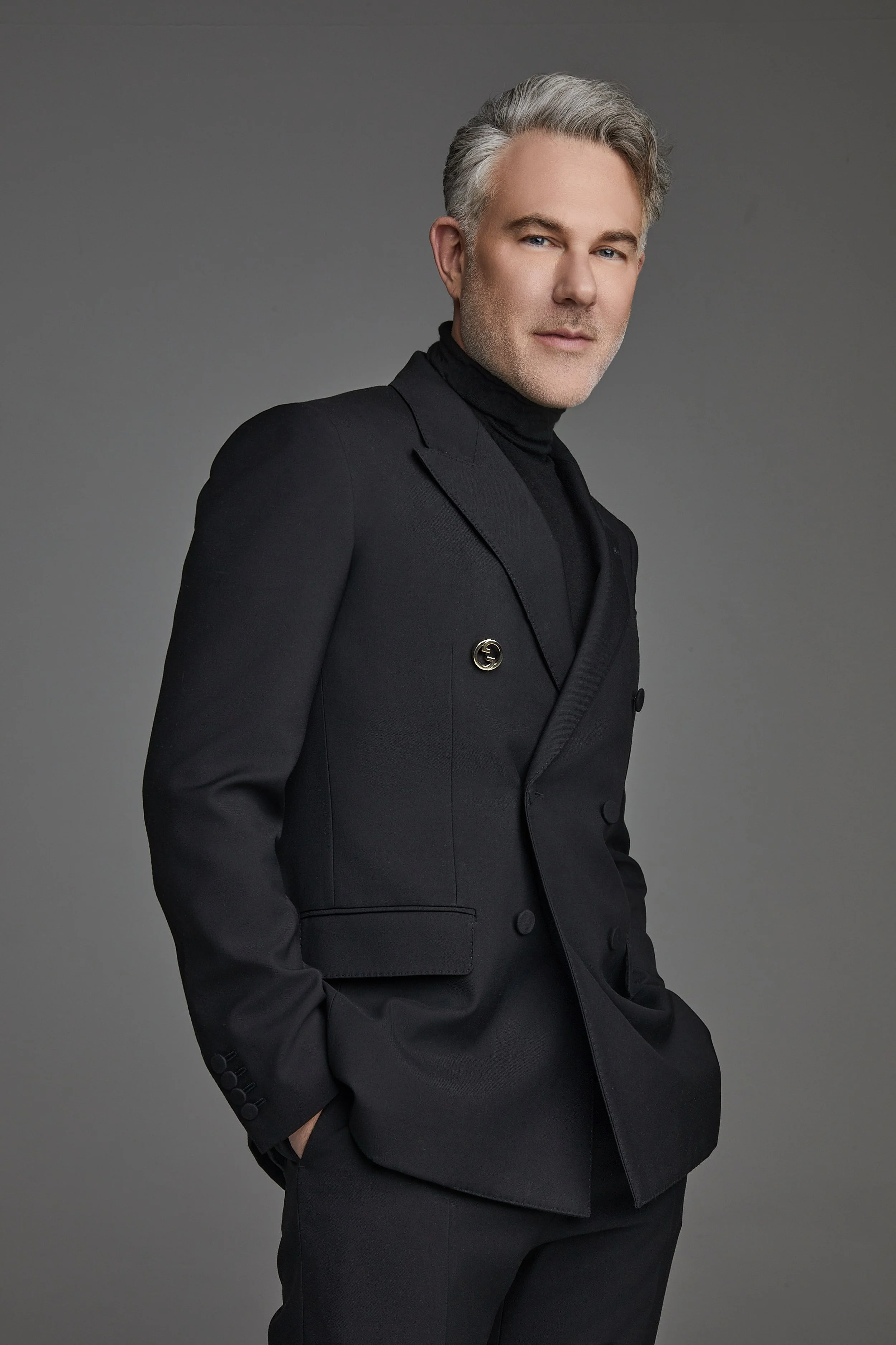 David Miskin wearing a Gucci black blazer, black turtleneck, and black pants, standing against a plain gray background, with his right hand in his pocket and looking at the camera.