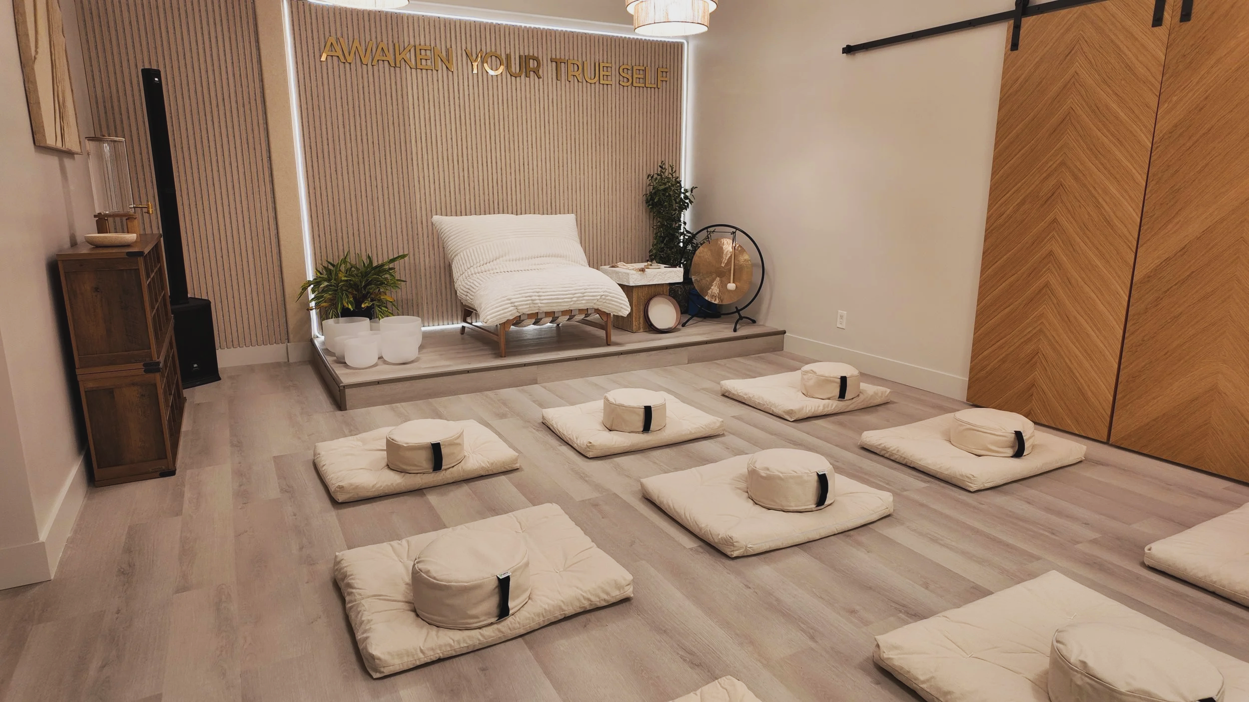 A peaceful meditation space with cushions and pillows on light wooden floor, a small elevated platform with a white blanket, plants, a gong, a singing bowl, and a banner that reads "Awaken Your True Self."
