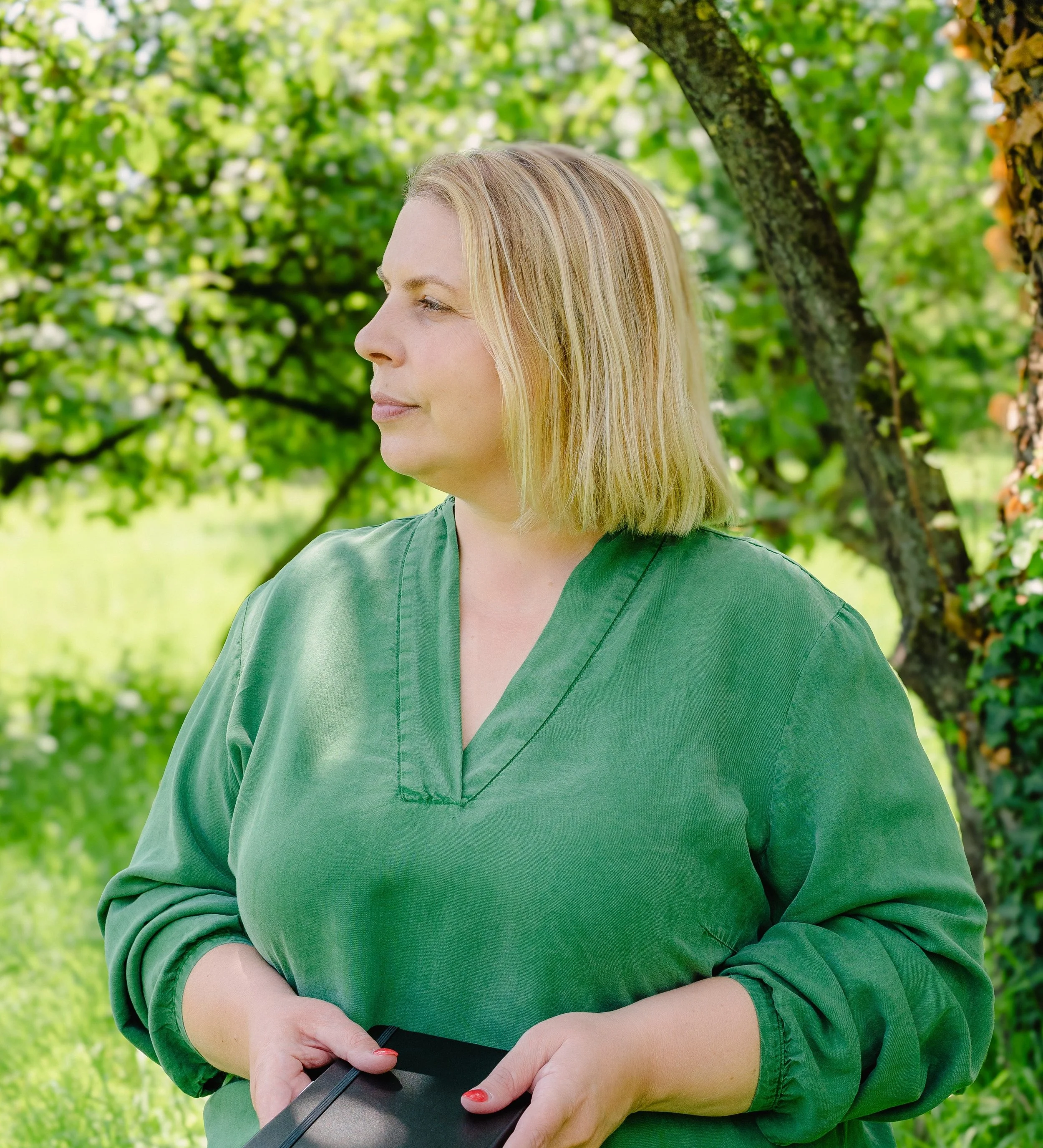 Elke Mertens, onafhankelijk archeologisch adviseur. Op de foto is ze herkenbaar als een vrouw met blond haar in een groene blouse. Ze staat buiten in een tuin, omringd door groene bladeren en bomen en houdt een notitieboekje vast.