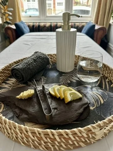 A wicker tray on a table with a soap dispenser, a glass of water, a rolled gray towel, a black serving plate with lemon wedges, and a pair of tongs, with a window and curtains in the background.