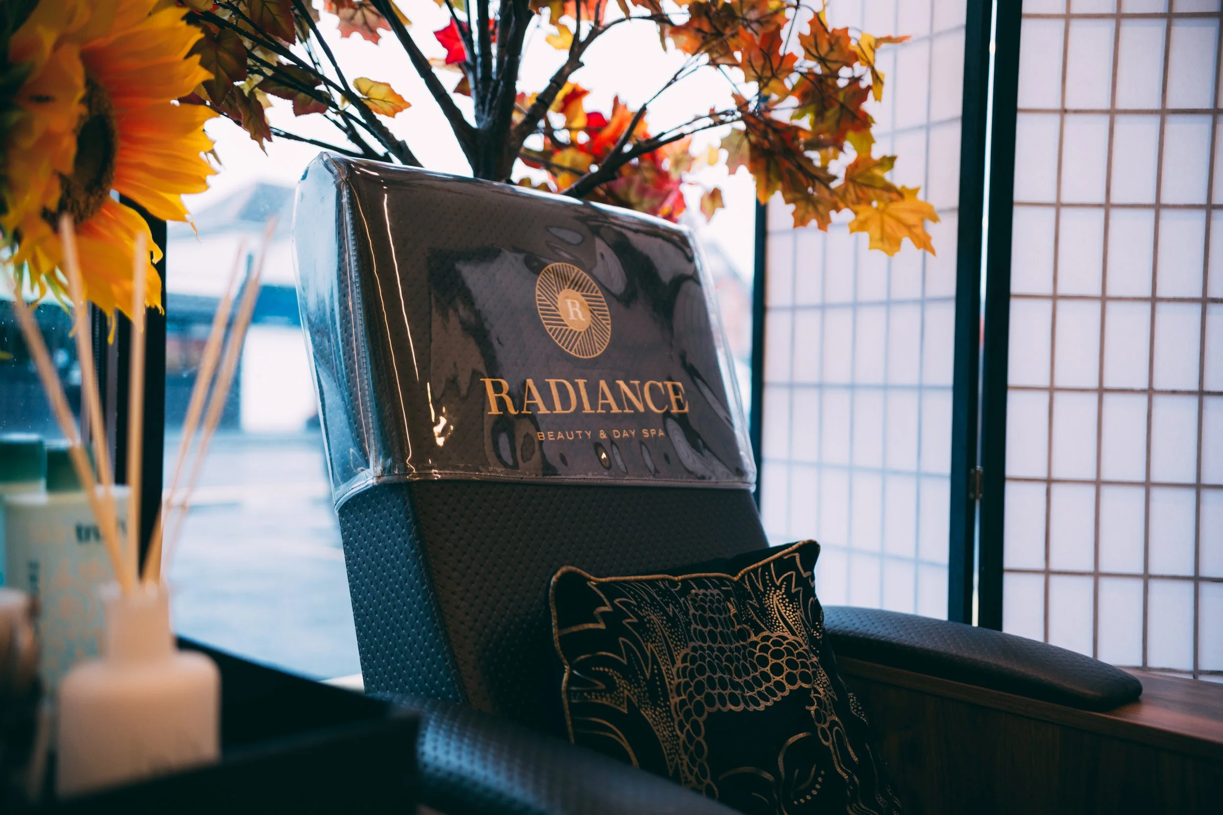 A chair with the word Radiance beauty and day spa on a sign, inside a space decorated with artificial autumn leaves, a sunflower, and a shoji screen, suggesting a spa or beauty salon setting.
