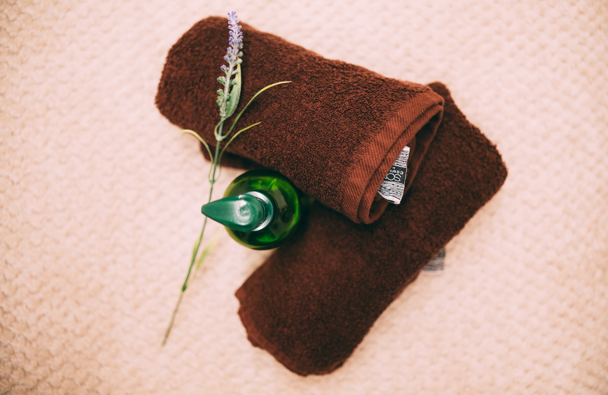 Two brown towels rolled with a small purple flower and a green soap dispenser placed on top, photographed on a beige textured surface.