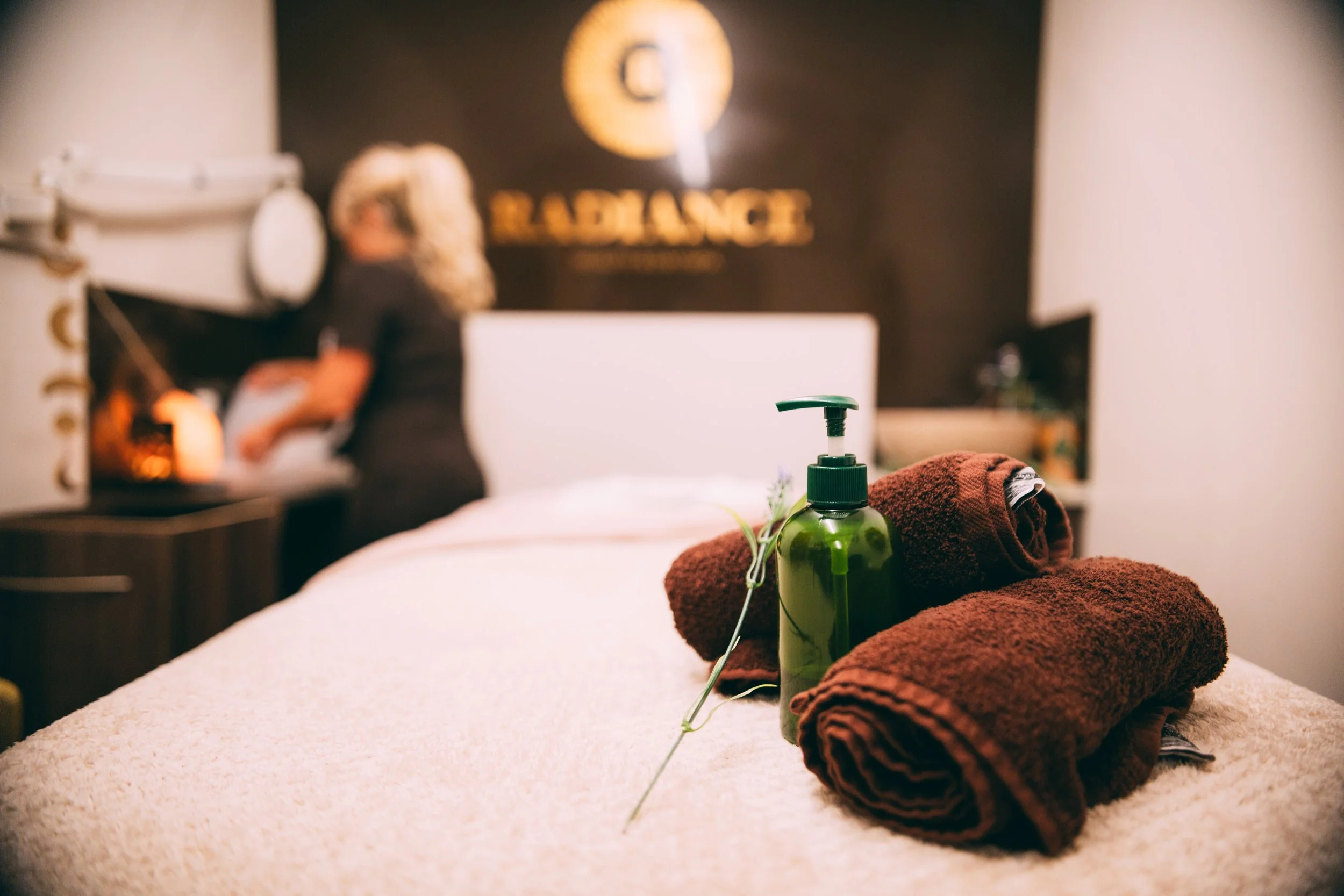 A hotel room with rolled brown towels, a green soap dispenser, and a small plant on the bed. Blurred background includes a woman near the reception desk and a sign reading 'RECEPTION' on the wall.