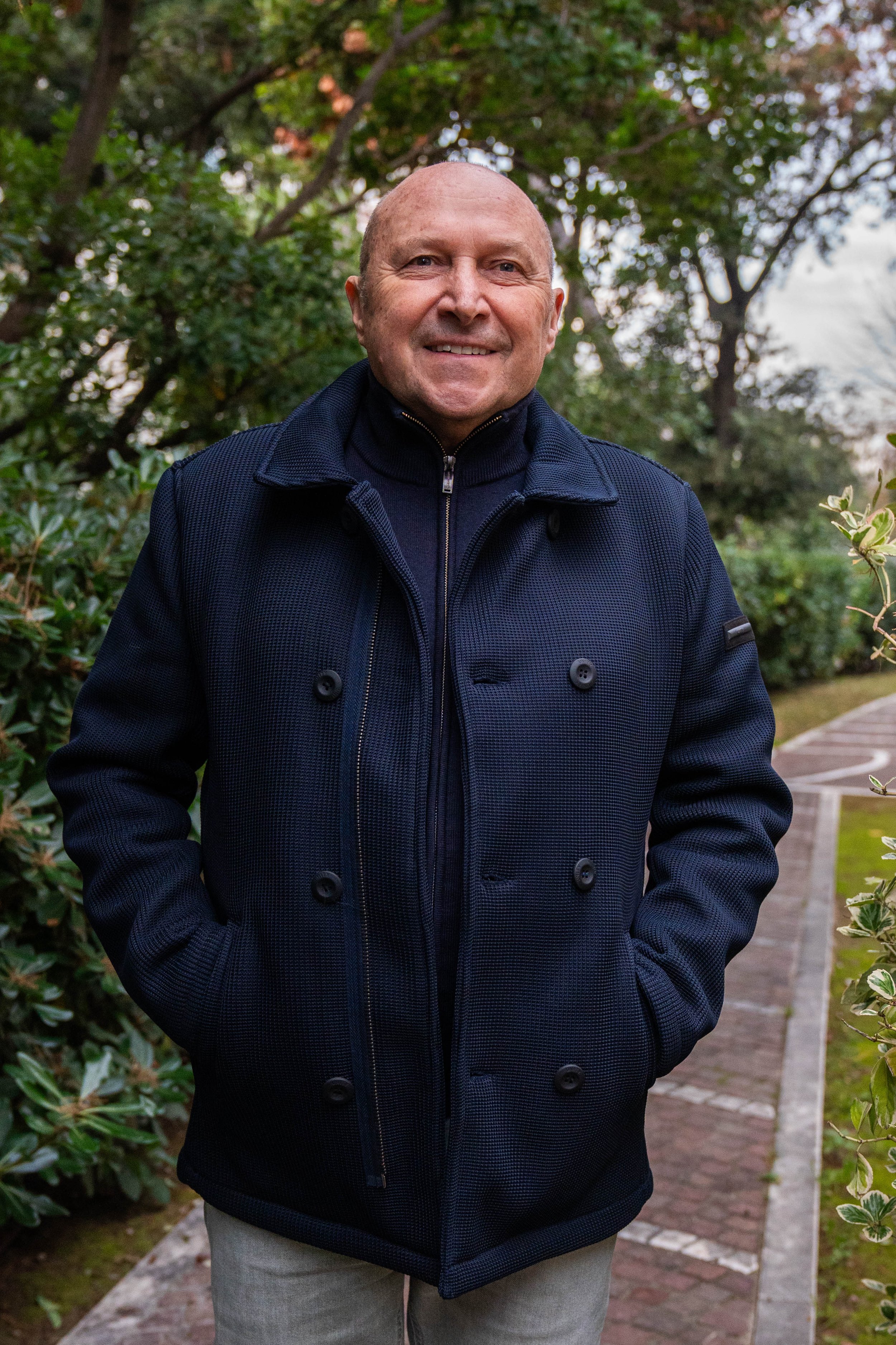 Uomo sorridente con sopracciglia sottili, cappotto blu scuro con tasche, in piedi in un giardino con alberi e cespugli.