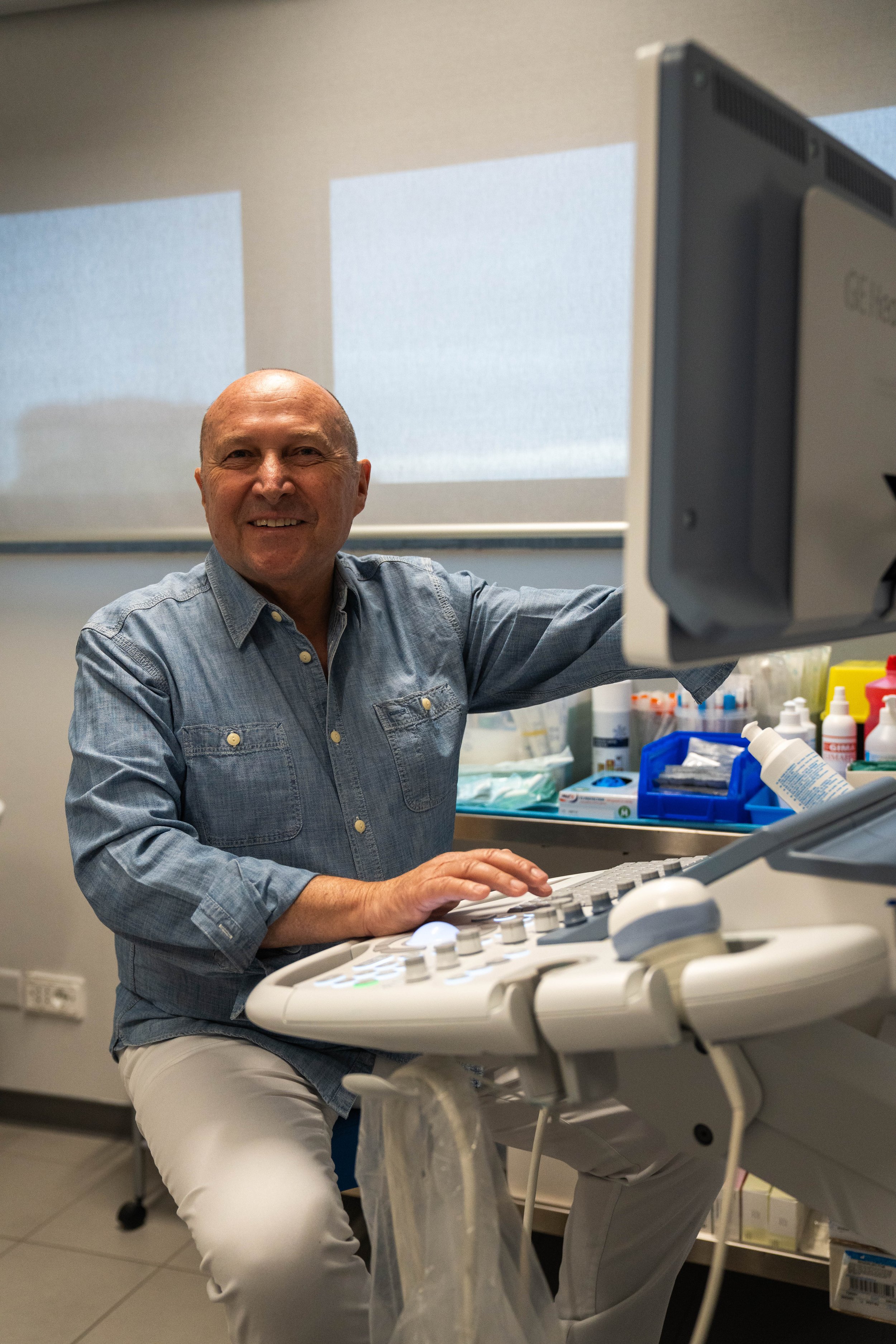 Uomo sorridente seduto vicino a un apparecchio medico, con strumenti e prodotti sanitari sullo sfondo in uno studio medico.