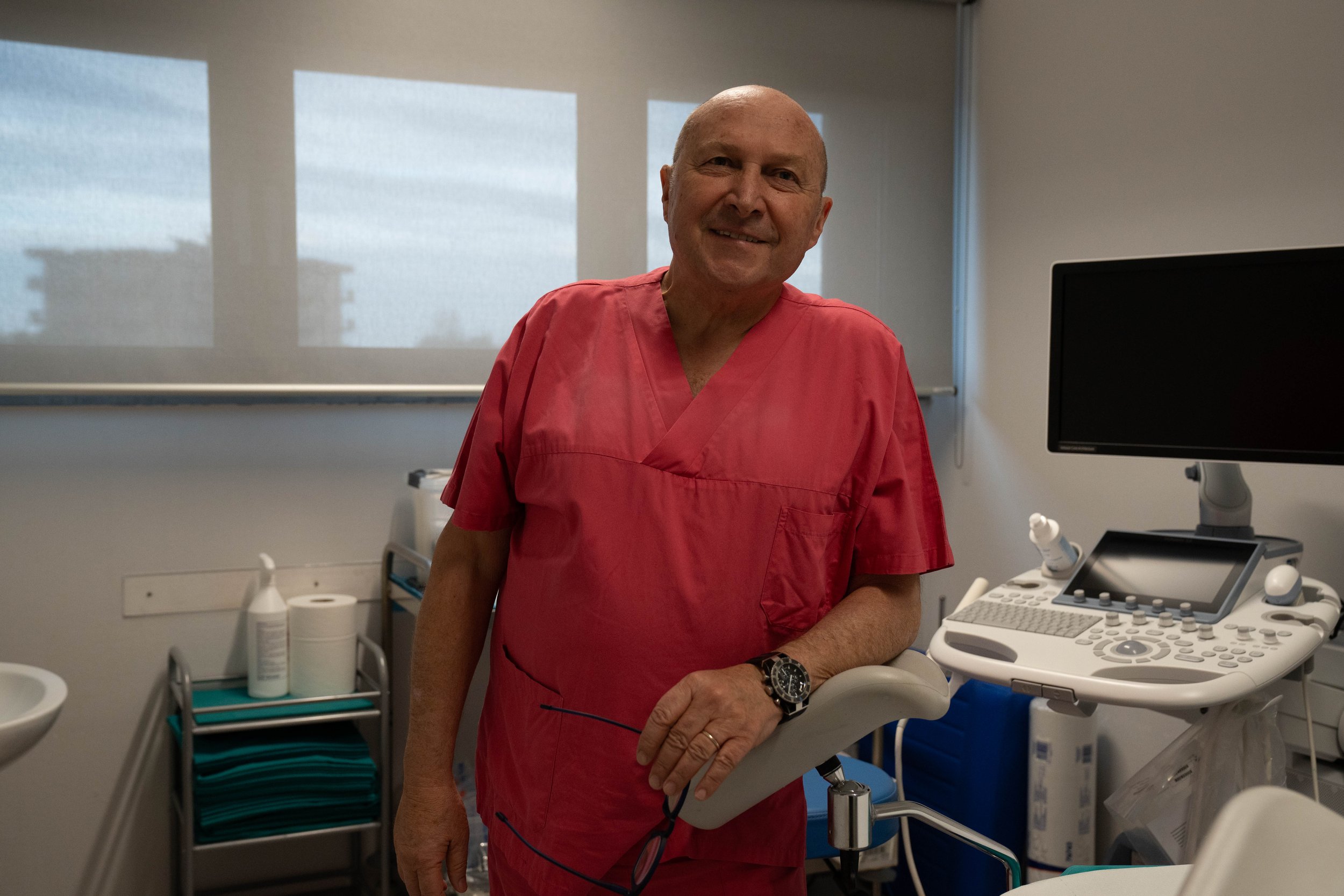 Un uomo in divisa da medico in uno studio medico o ambulatorio, con apparecchiature mediche e computer sullo sfondo.
