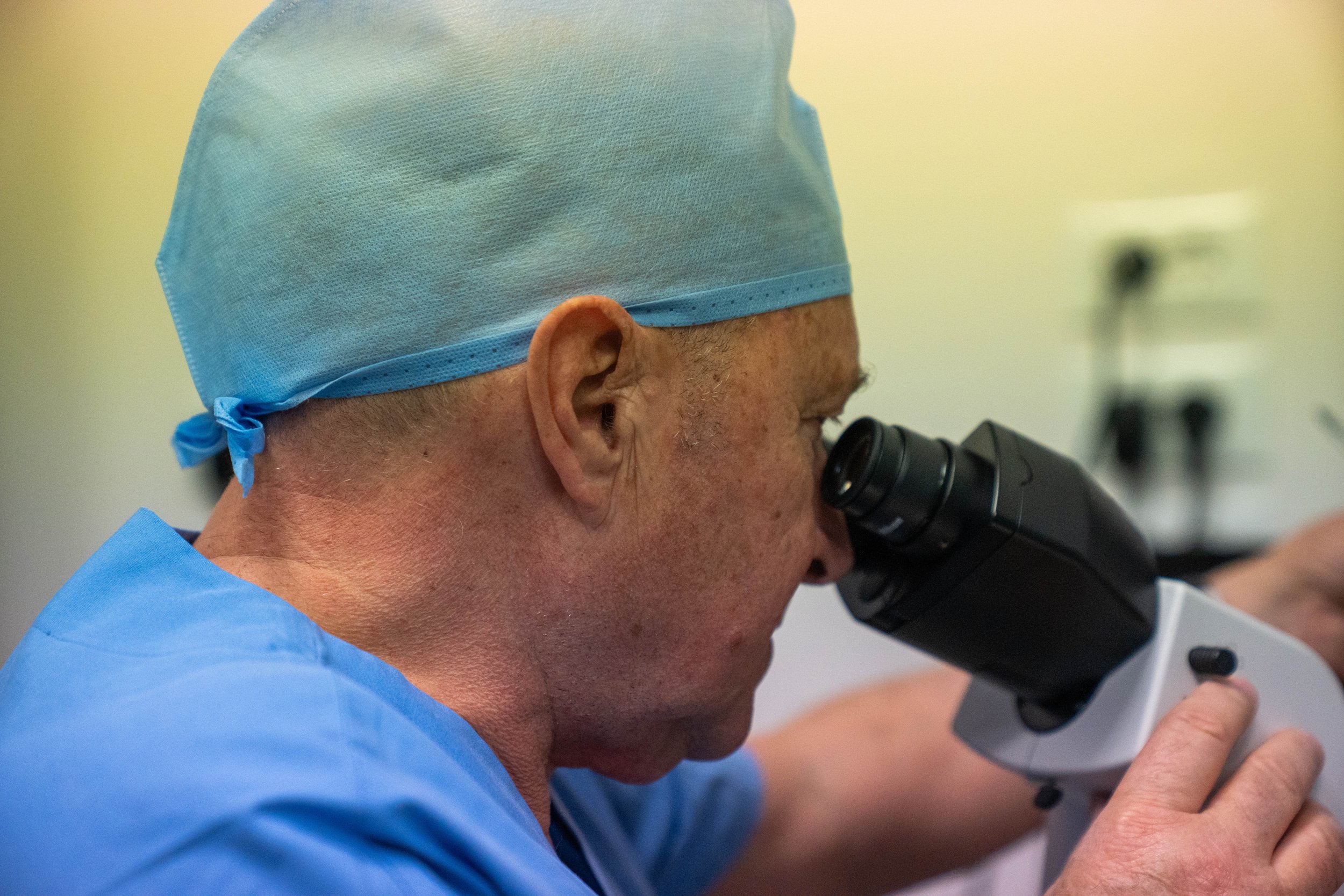 Un medico indossa un camice blu, un cappello chirurgico azzurro e utilizza un microscopio.