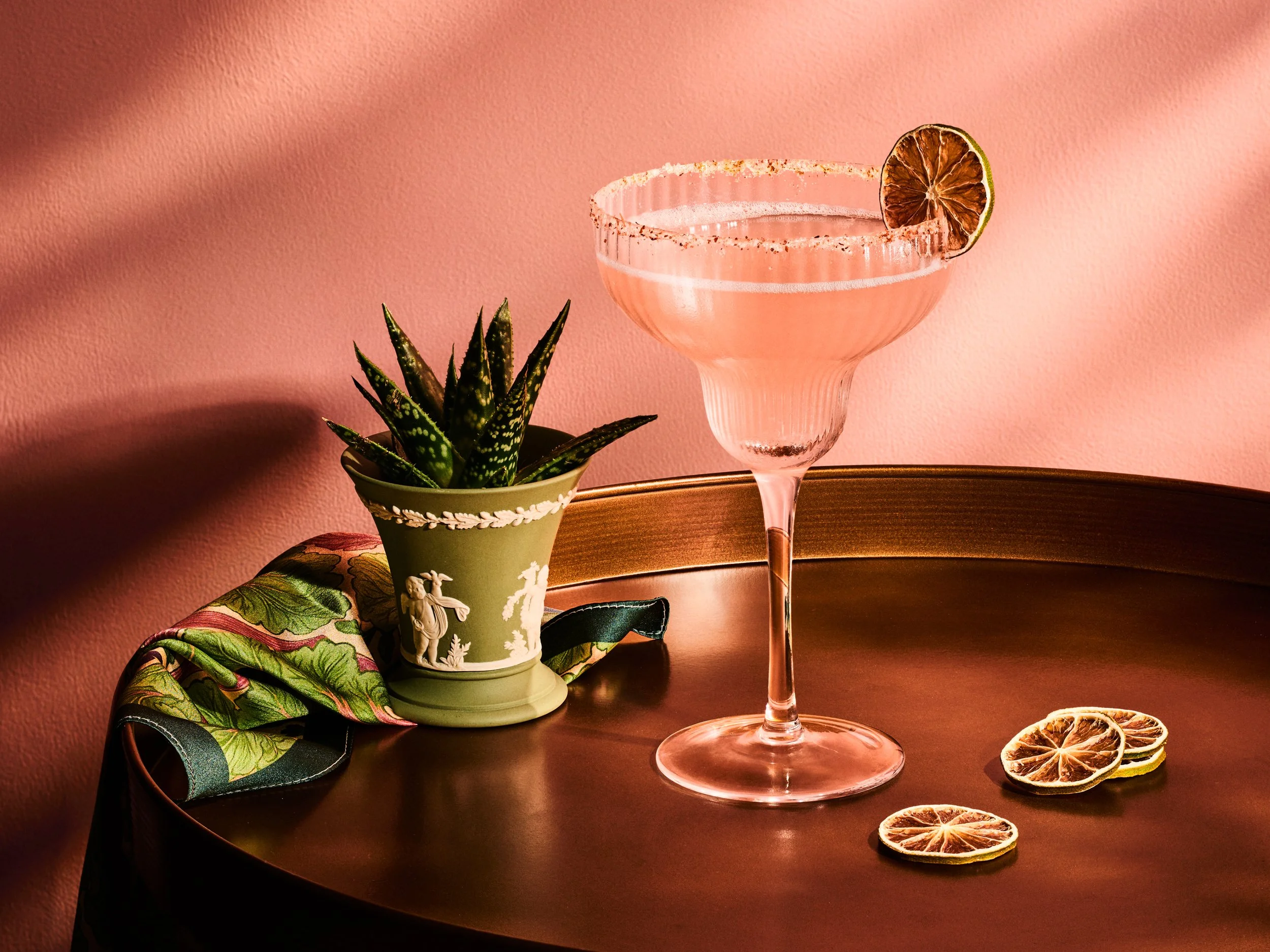 A pink cocktail in a margarita glass with a salted rim and dried lime slice garnish, placed on a dark wooden table. Next to the drink, there's a small potted succulent plant in a decorative green pot, and three dried lemon slices on the table. The background is pink with soft lighting creating shadows.