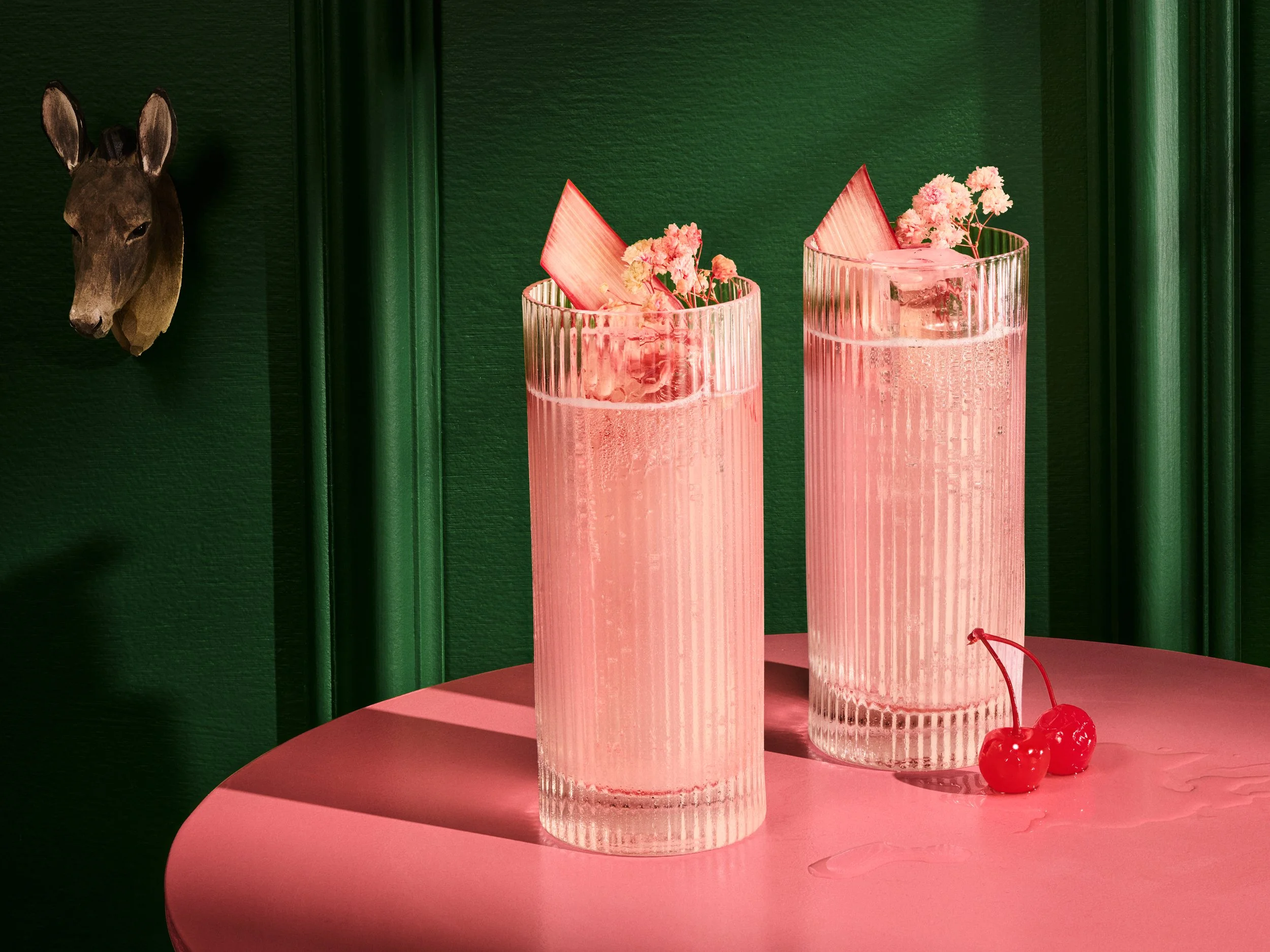 Two tall pink drinks in ribbed glasses garnished with pink flowers and pink paper decorations on a pink table, with two cherries placed near them.