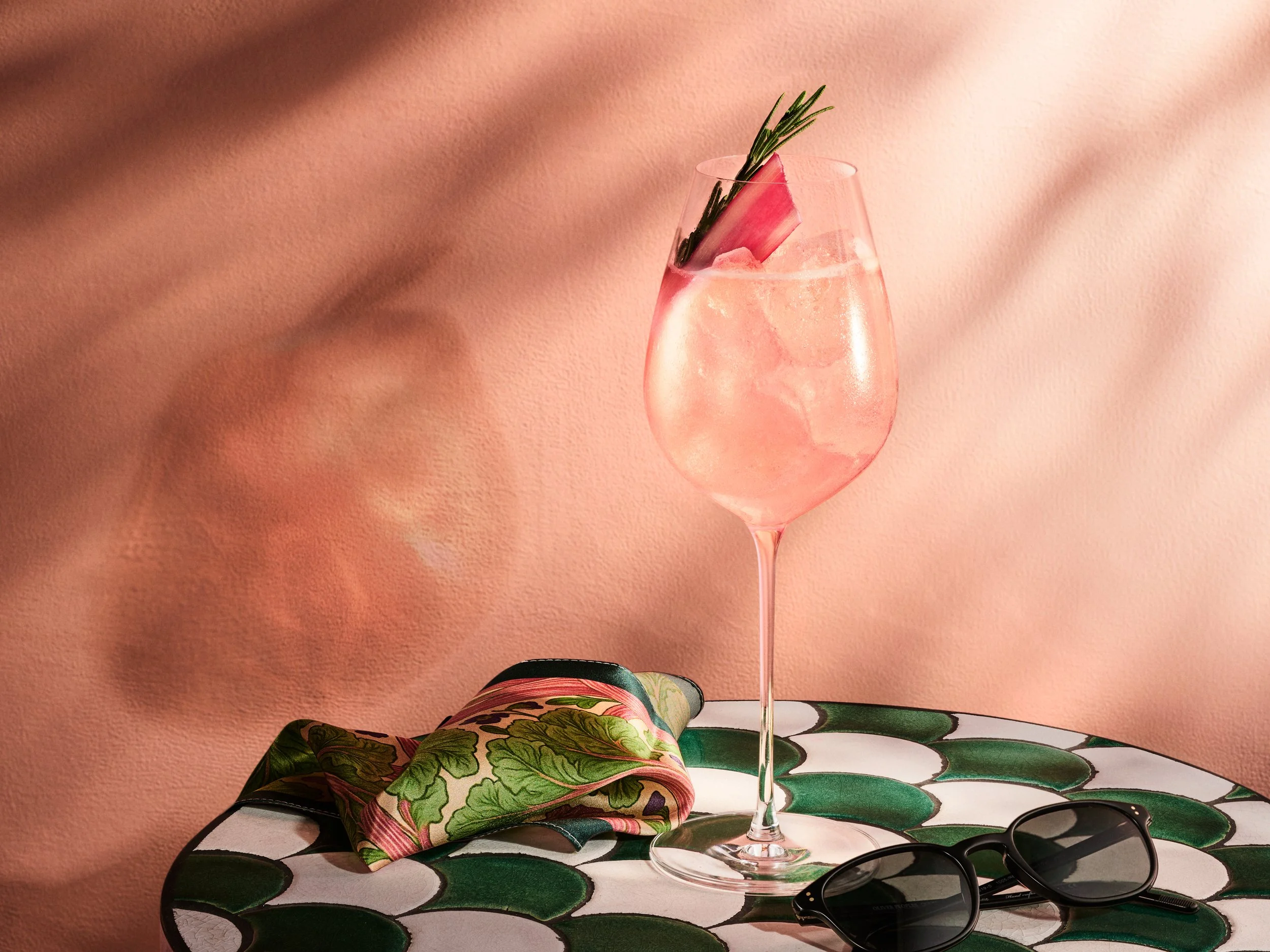 A pink cocktail in a tall wine glass garnished with a rosemary sprig and a piece of watermelon rind, on a patterned table with sunglasses and a colorful scarf, against a soft pink background.