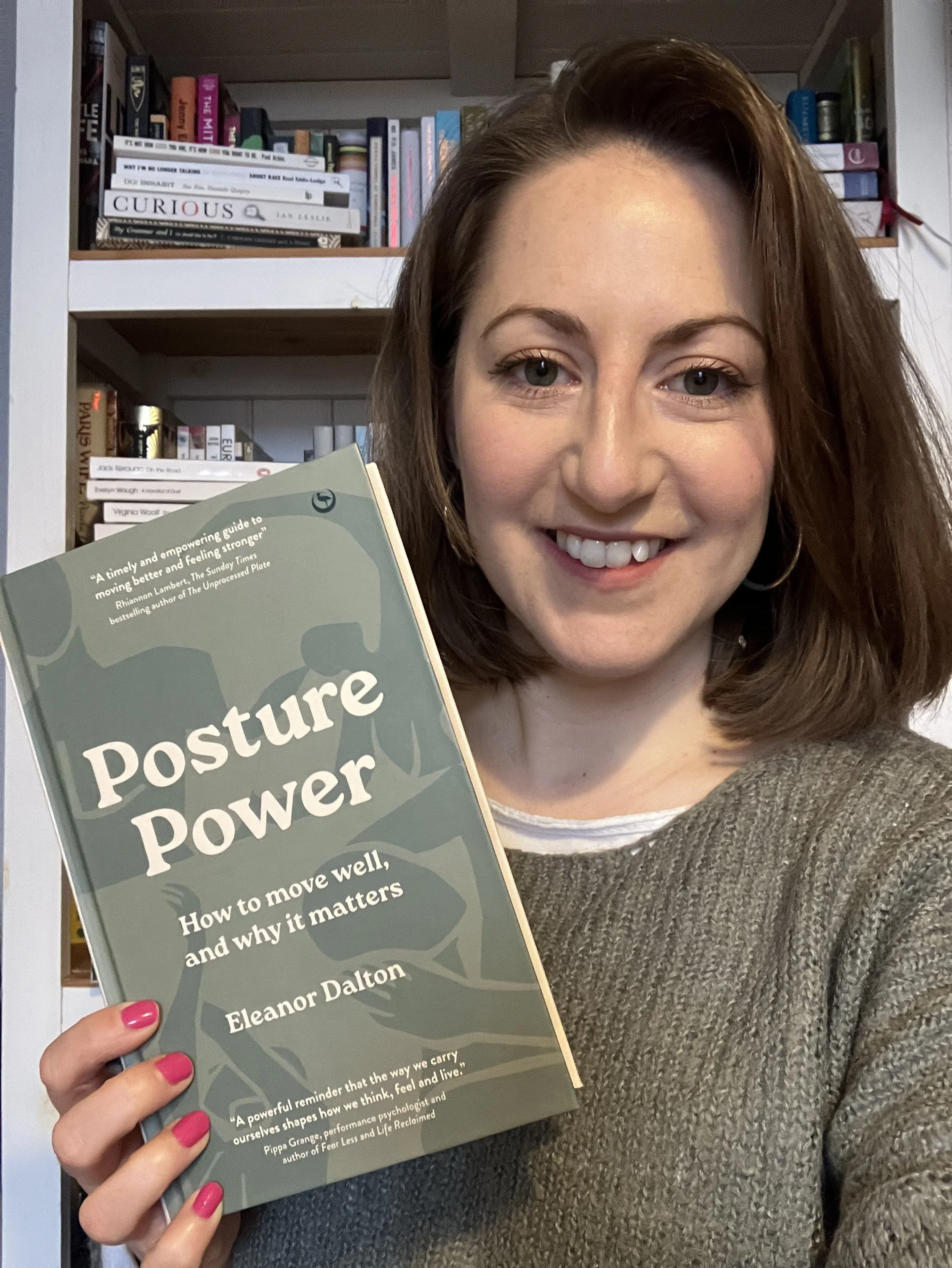 A woman with brown hair, smiling, holding a book titled 'Posture Power' in front of a bookshelf filled with various books.