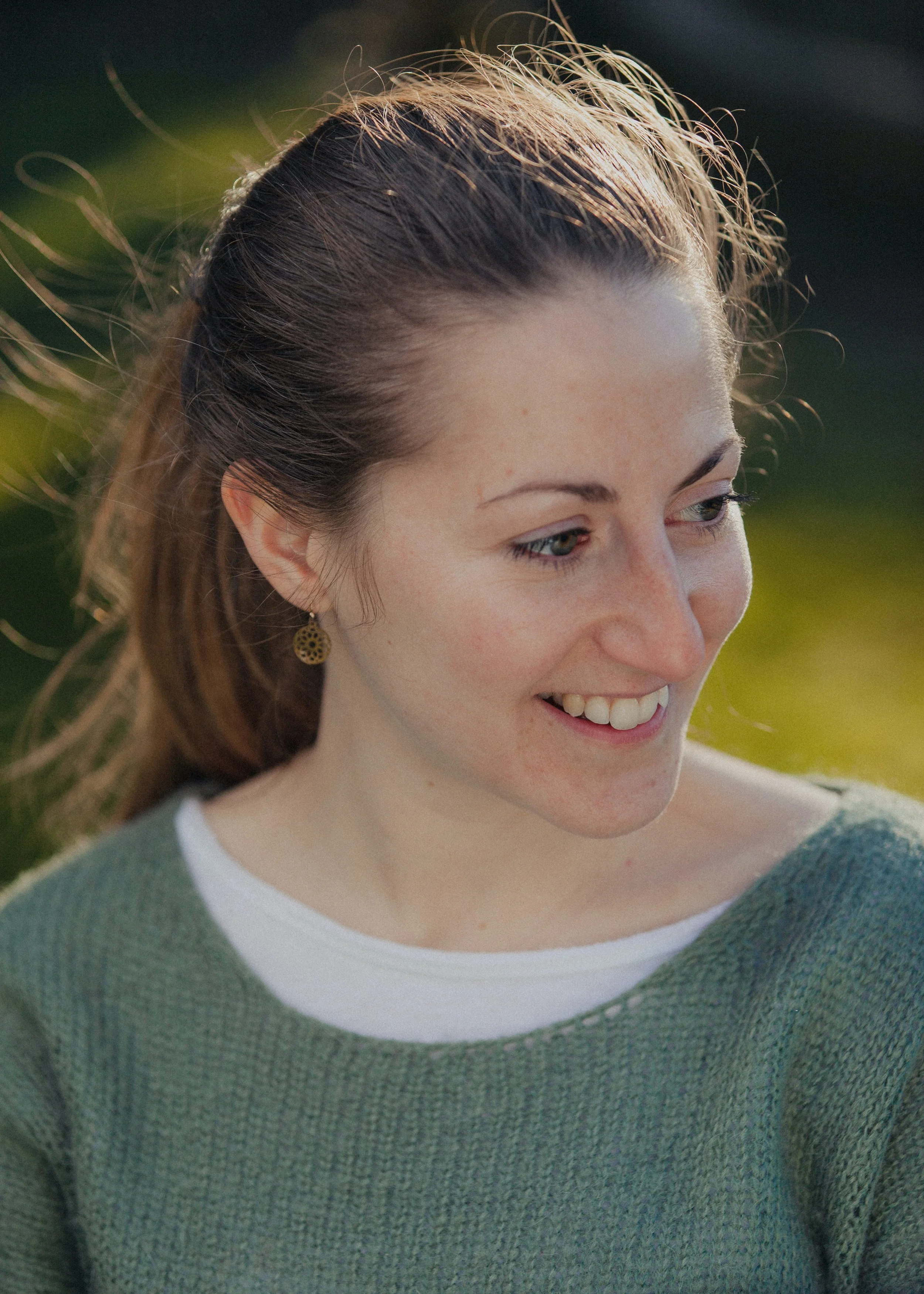 Close-up of a young woman smiling outdoors, wearing a green sweater and a white shirt underneath, with her hair blowing in the wind, and wearing a gold earring.