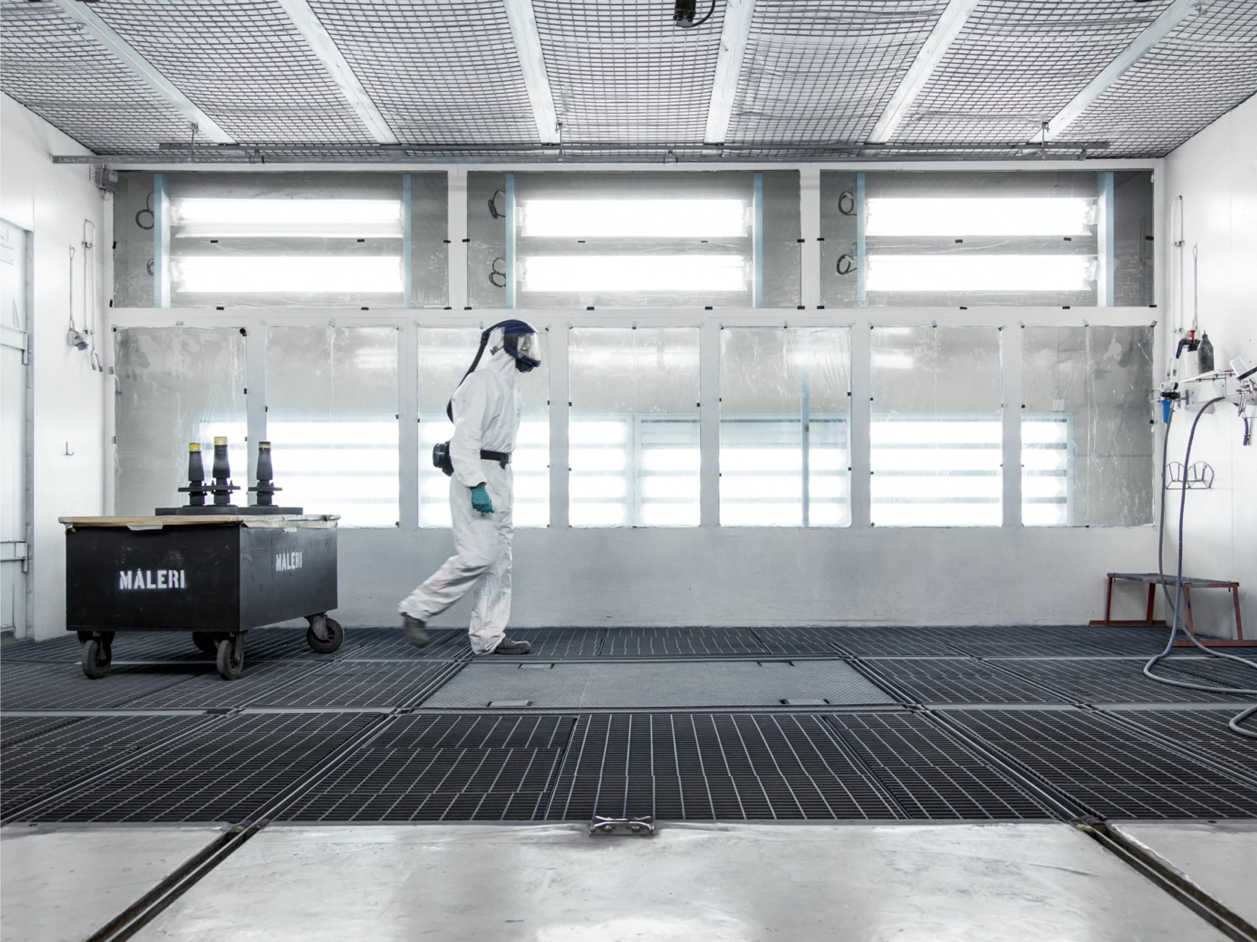 A person in protective gear walking inside a cleanroom or laboratory with a black cart labeled 'Maleri' and various equipment on the walls.