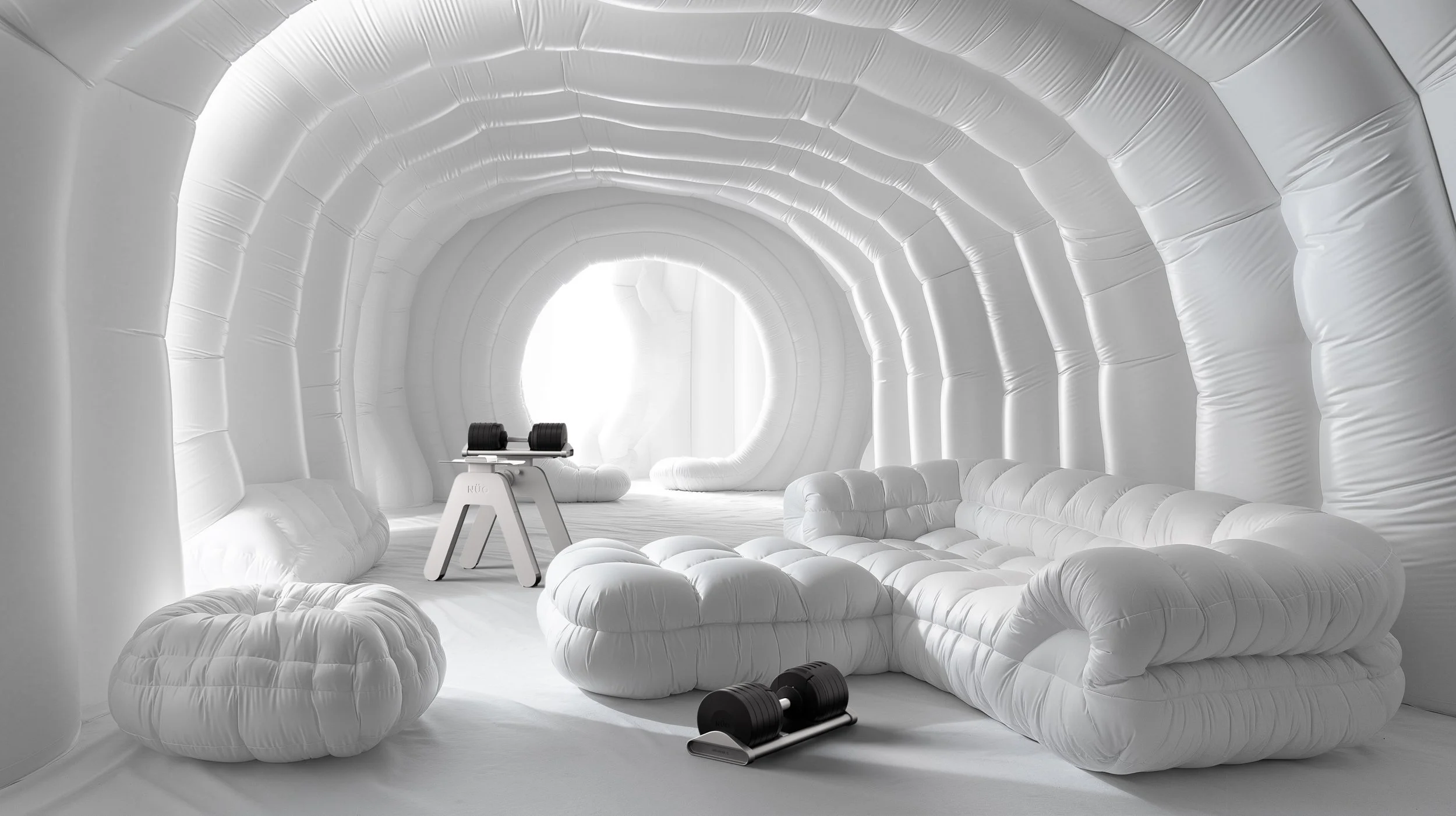 Interior of a futuristic, inflatable lounge with plush, white, cushioned furniture, and a set of black dumbbells on a white stand, all within an arched, white inflatable tunnel.