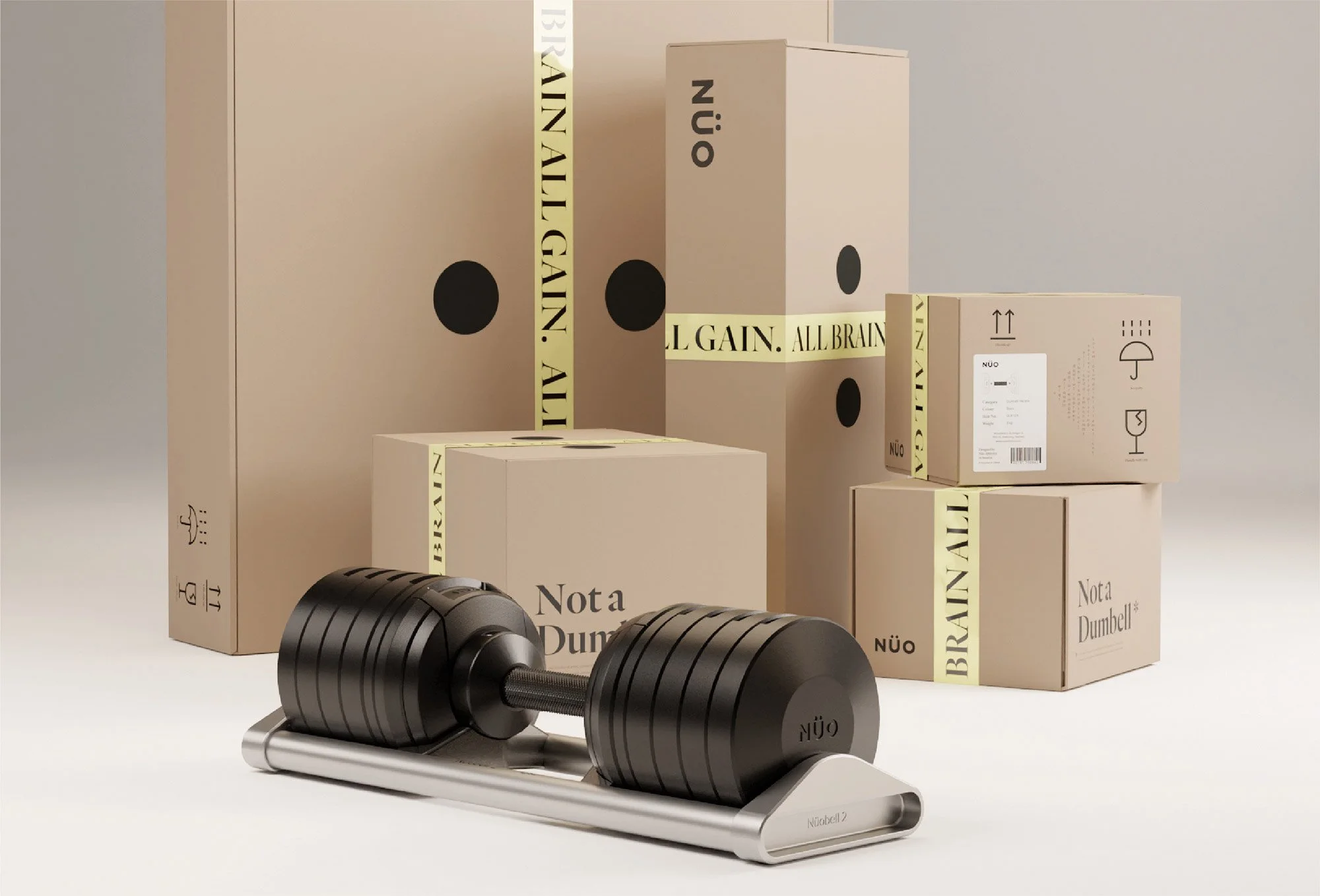 A pair of black dumbbells on a silver stand in front of beige boxes with black and yellow text and symbols, including the words 'Nota Dumbbell,' 'NÜO,' and 'ALL BRAIN.' The boxes are stacked and arranged on a plain light background.