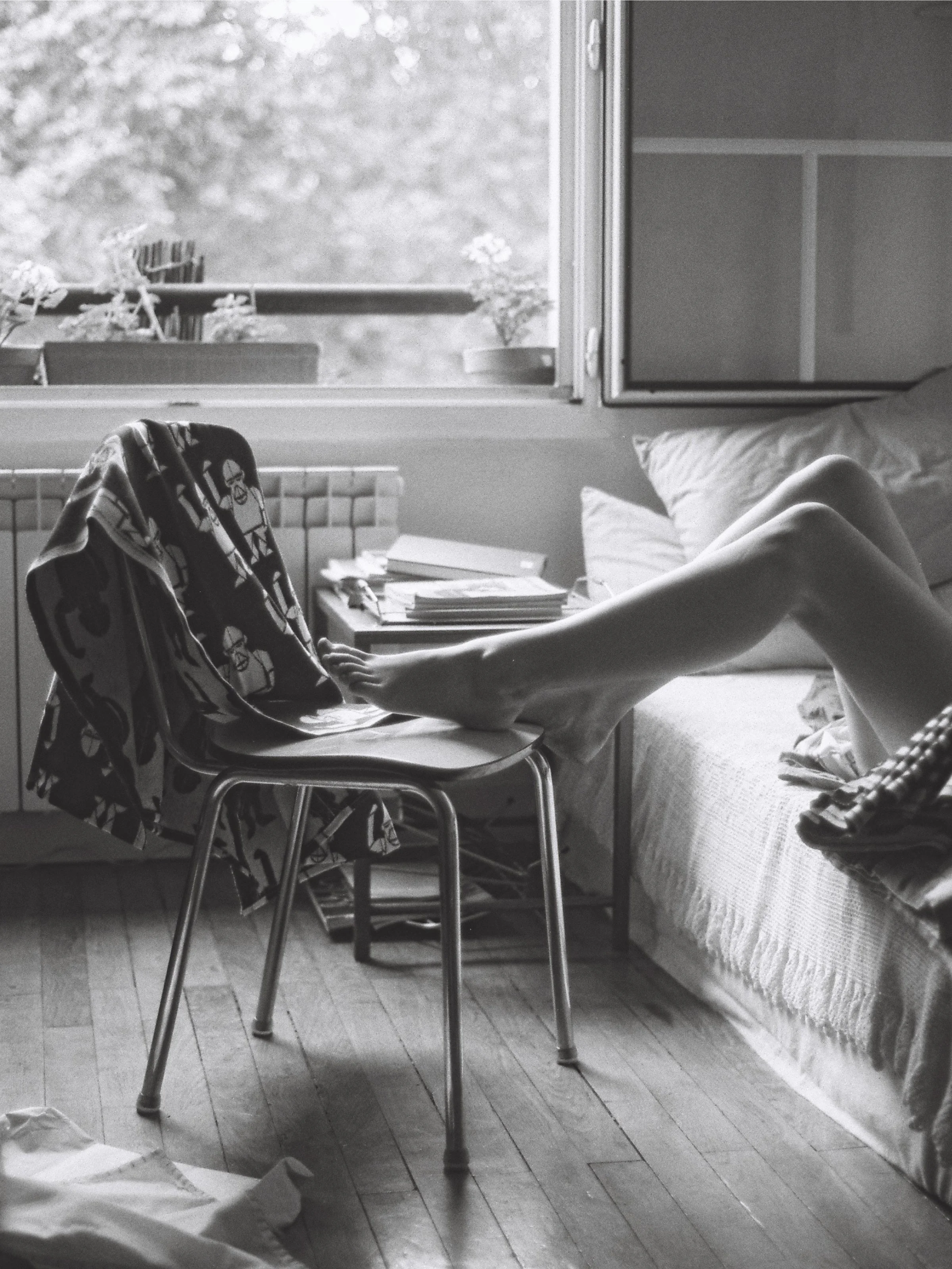 Black and white photo of a person lying on a bed with their legs extended onto a chair, with their feet resting on the chair's seat. An open window with plants on the windowsill and a bookshelf is visible in the background.
