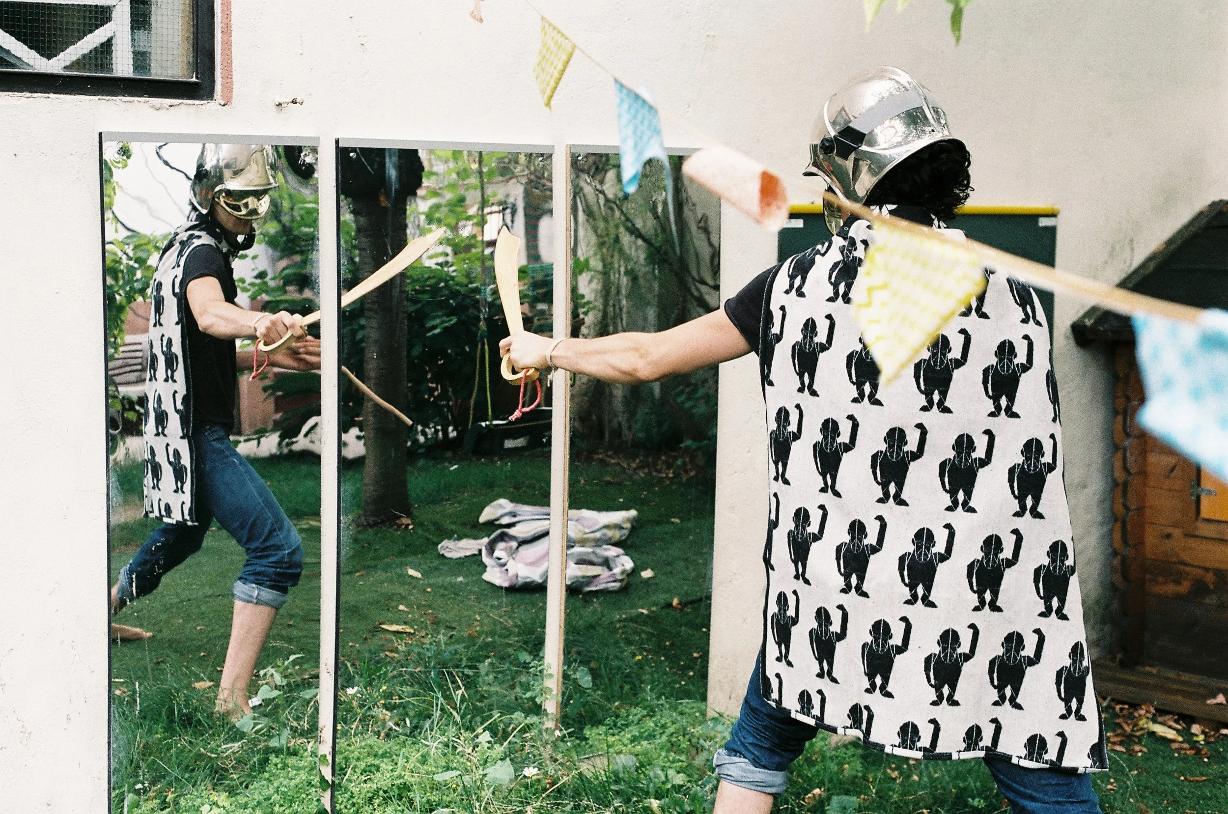 A person wearing a black t-shirt with a dog pattern, scissors in hand, through a mirror, with a mirror's reflection showing them holding a pair of scissors and a mirror in an outdoor area with grass, trees, and outdoor chairs, adorned with colorful bunting.