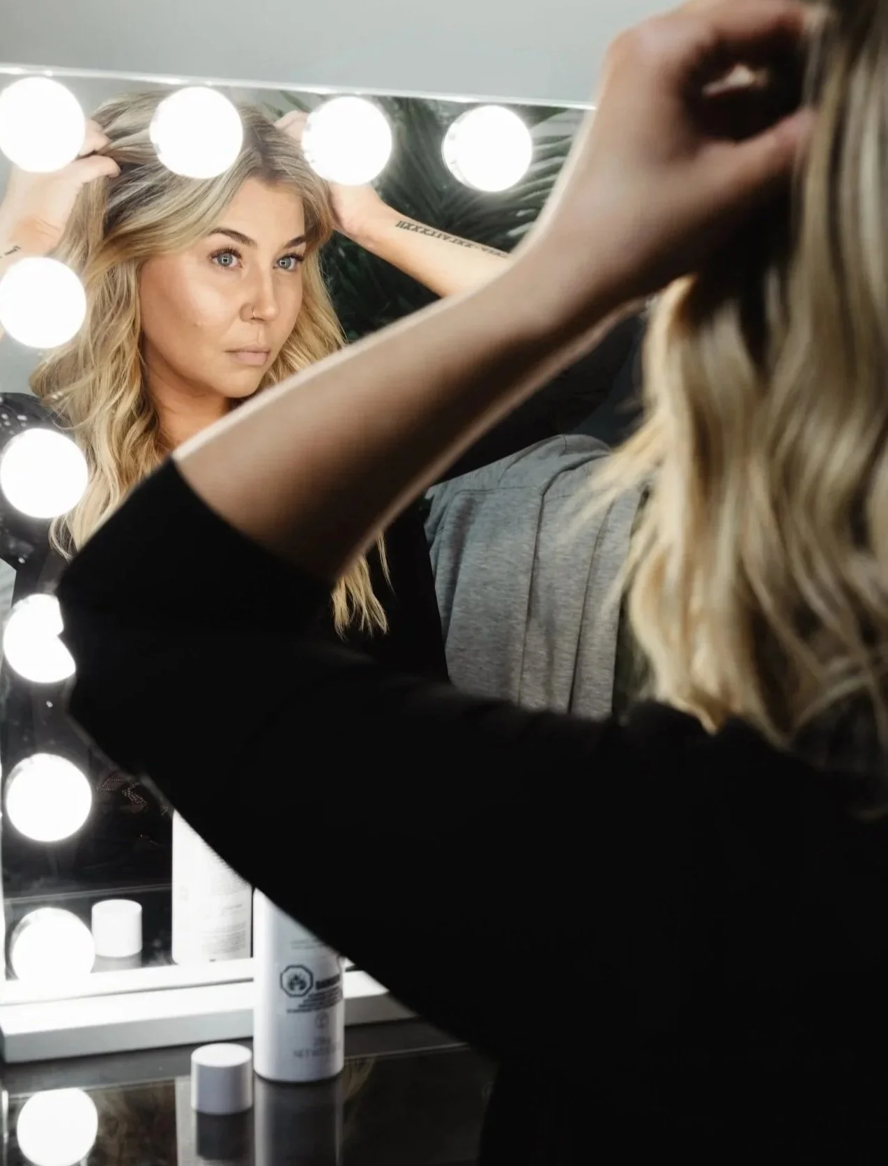 A woman with blonde hair looking at her reflection in a lit makeup mirror, adjusting her hair.