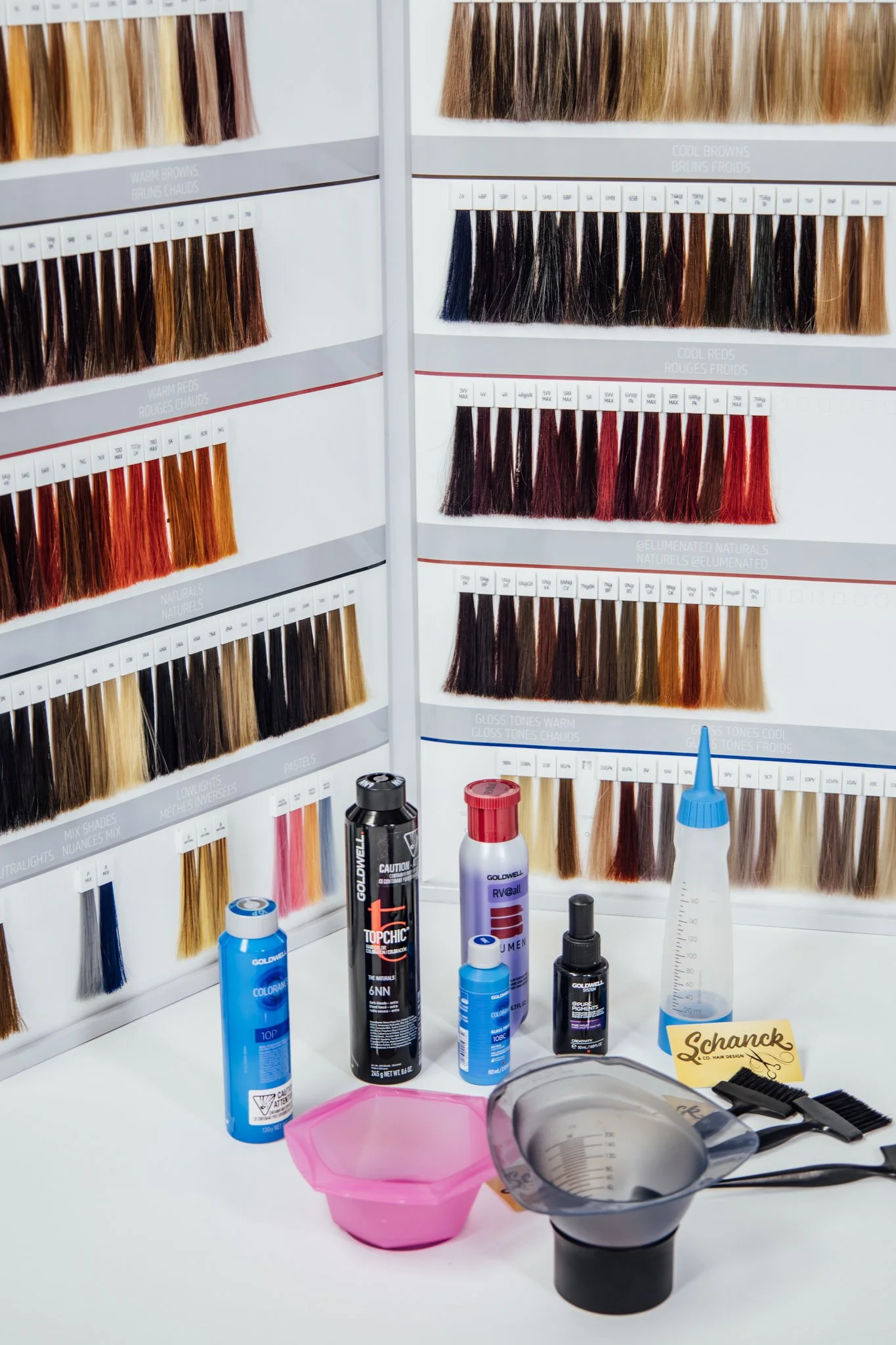 Display of hair color samples arranged in rows, with various hair dye bottles, a spray bottle, mixing bowl, brushes, and a card on a white table.