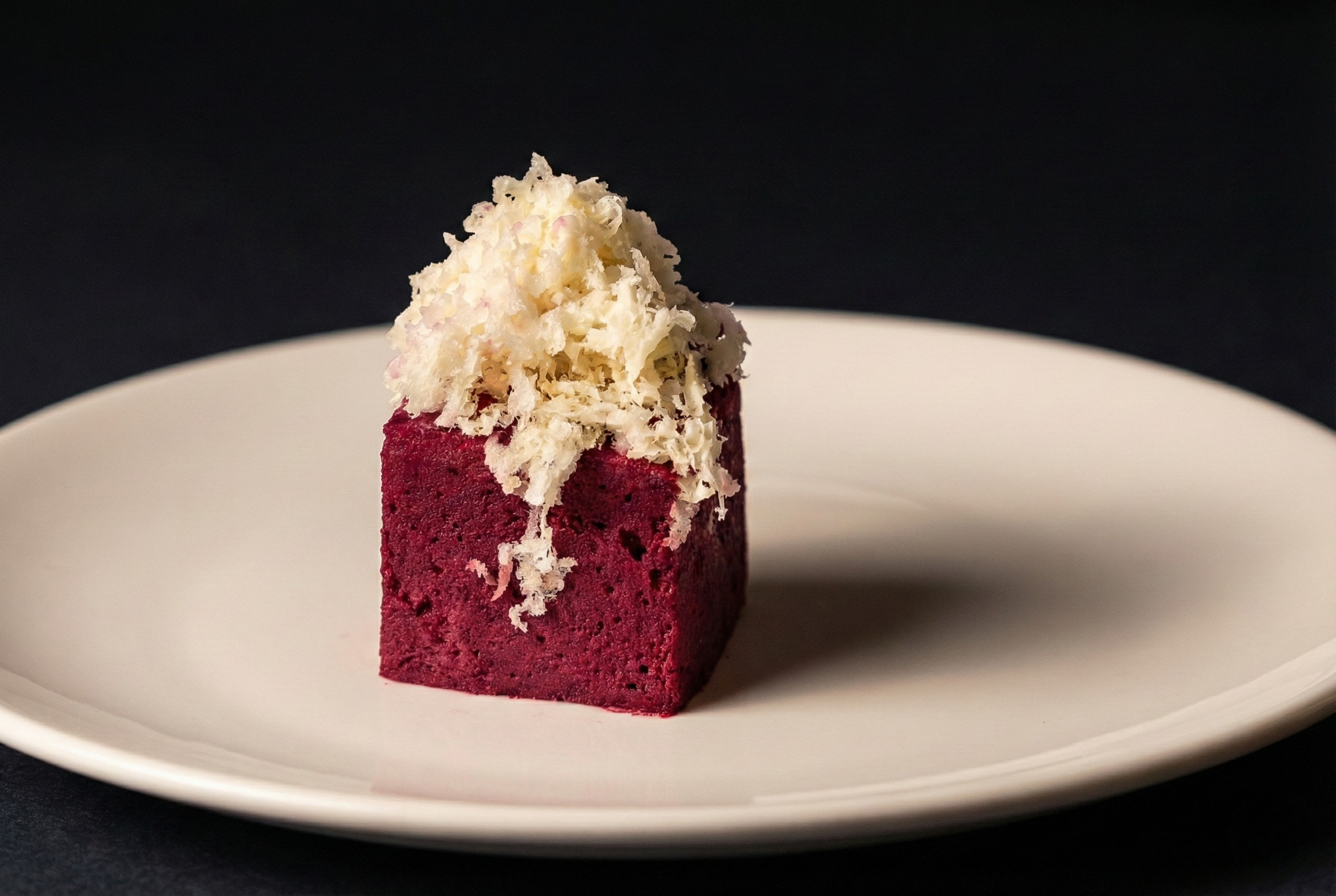 Ein rotes Dessert mit weißer Schlagsahne und Schokoladenraspeln auf einem weißen Teller vor schwarzem Hintergrund.