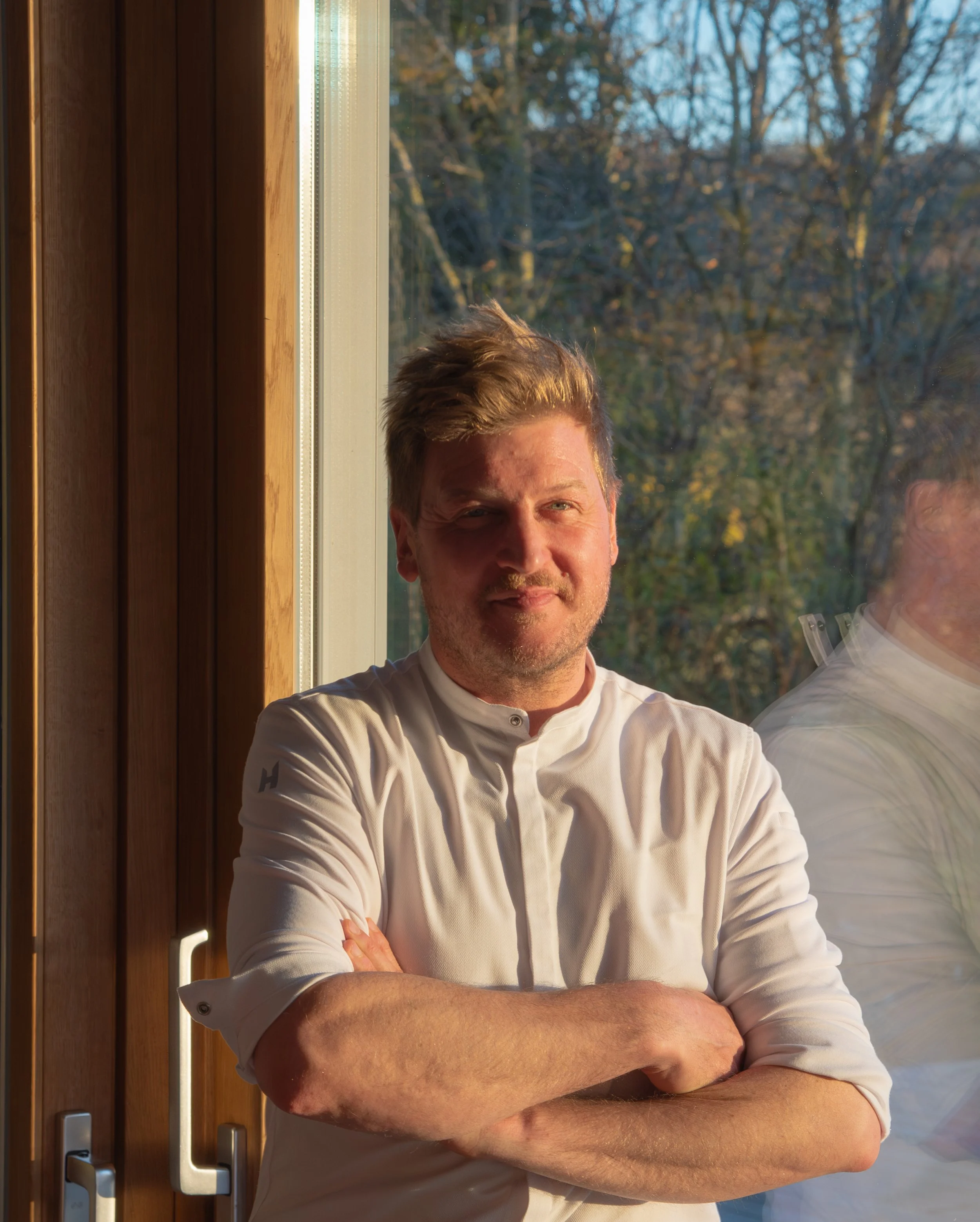 Mann mit weißen Kochjacke steht vor einem Fenster, Sonnenlicht beleuchtet sein Gesicht, im Hintergrund Bäume
