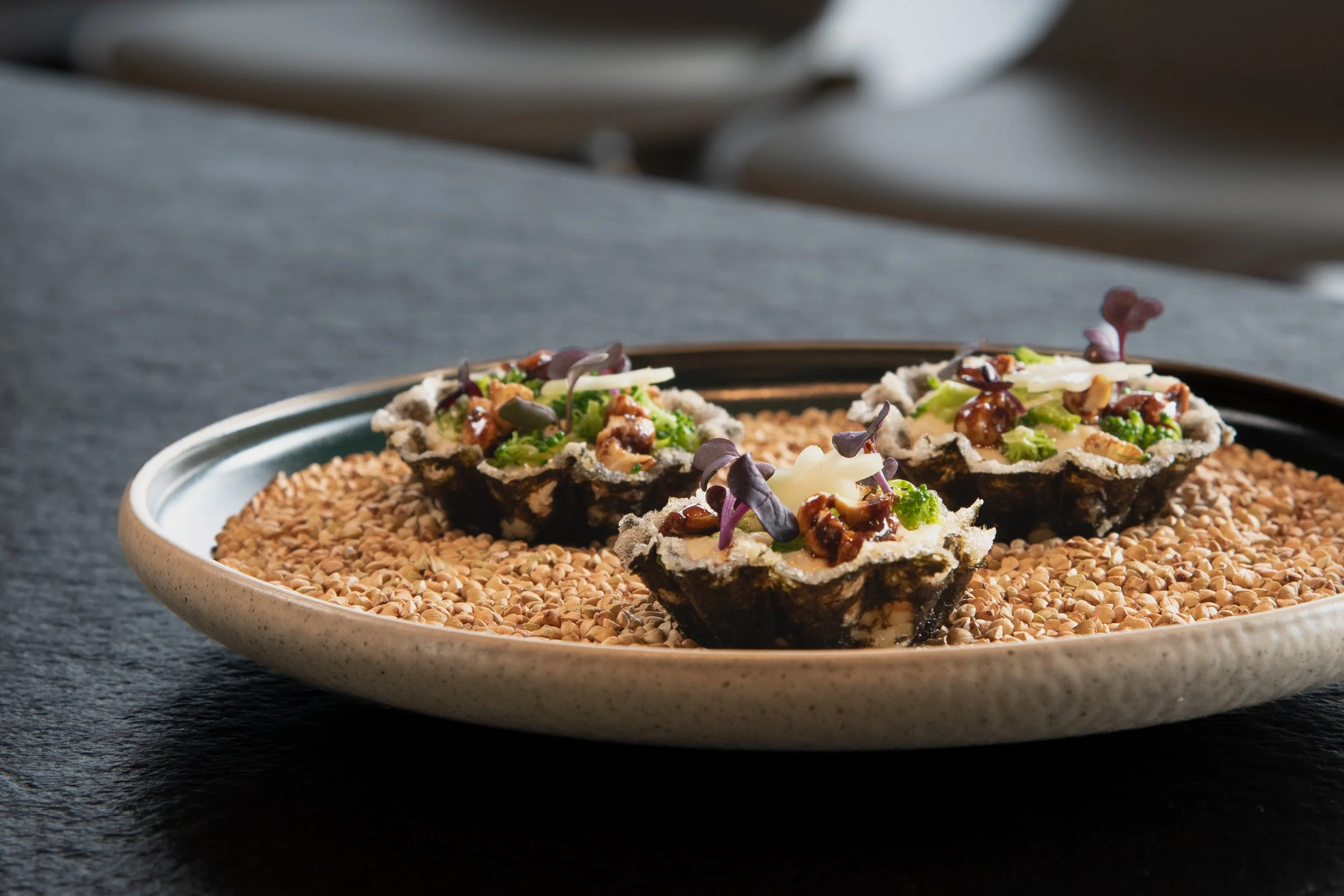Drei schwarze Tartlet mit grünen und weißen Zutaten, auf einem Teller mit Sesamsamen, auf einem dunklen Tisch