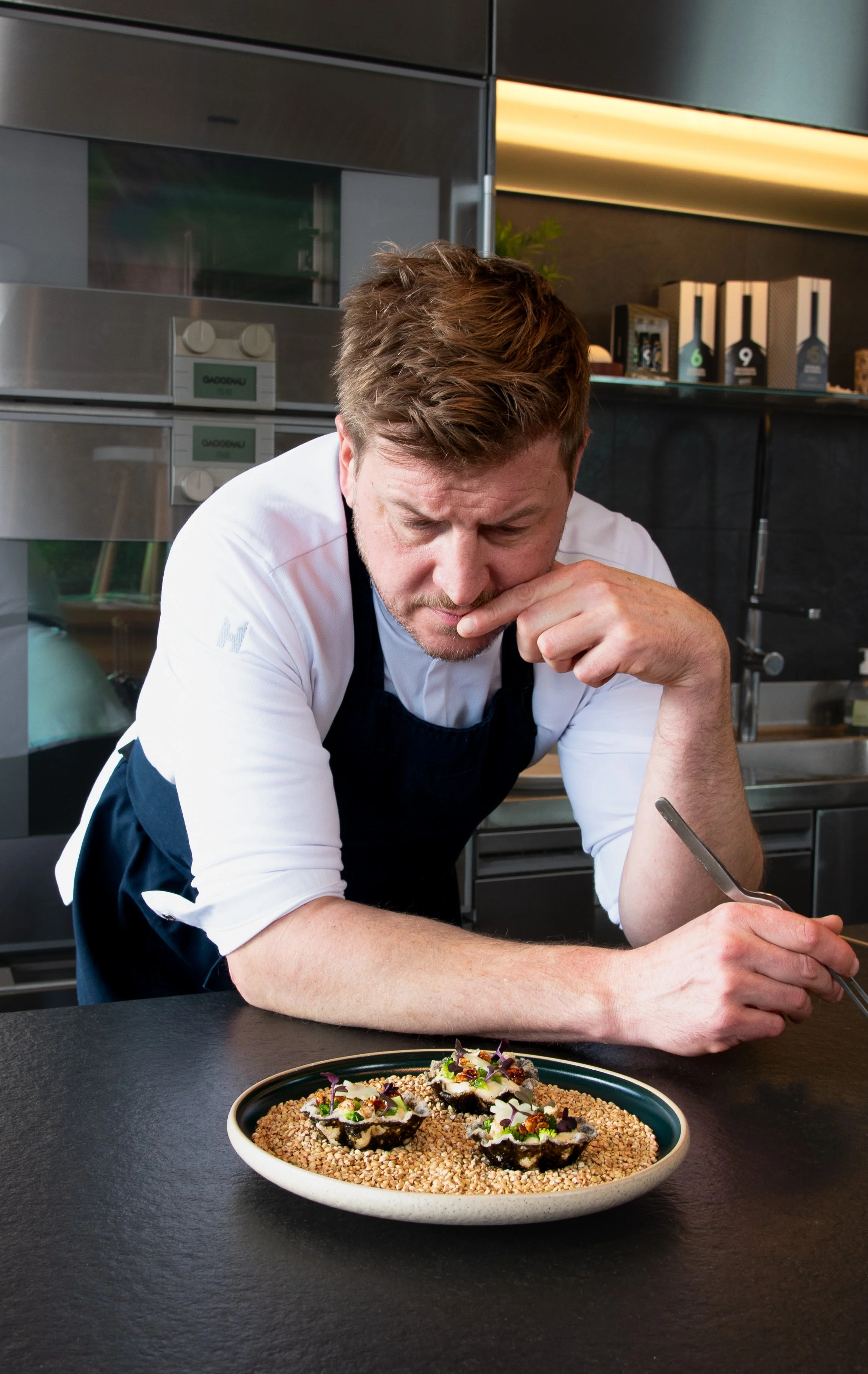 Ein Koch mit braunem Haar und Bart, in weißem Hemd und schwarzer Schürze, arbeitet in einer modernen, dunklen Küche, während er dekorative Gerichte auf einem schwarzen Tisch präsentiert.