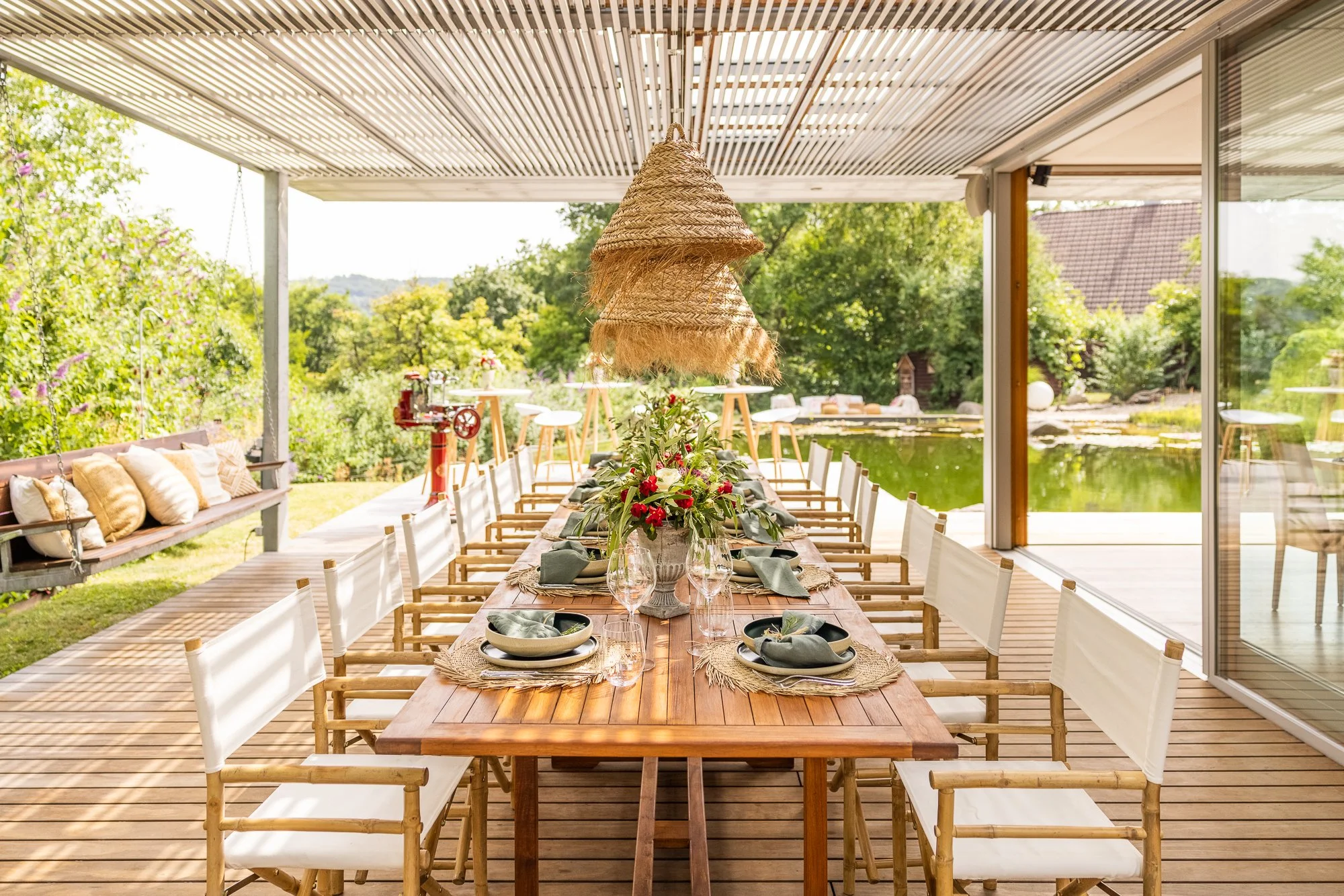 Ein festlich gedeckter Gartentisch mit Blumen und Geschirr auf einer Terrasse mit Blick auf einen See und grüne Bäume.