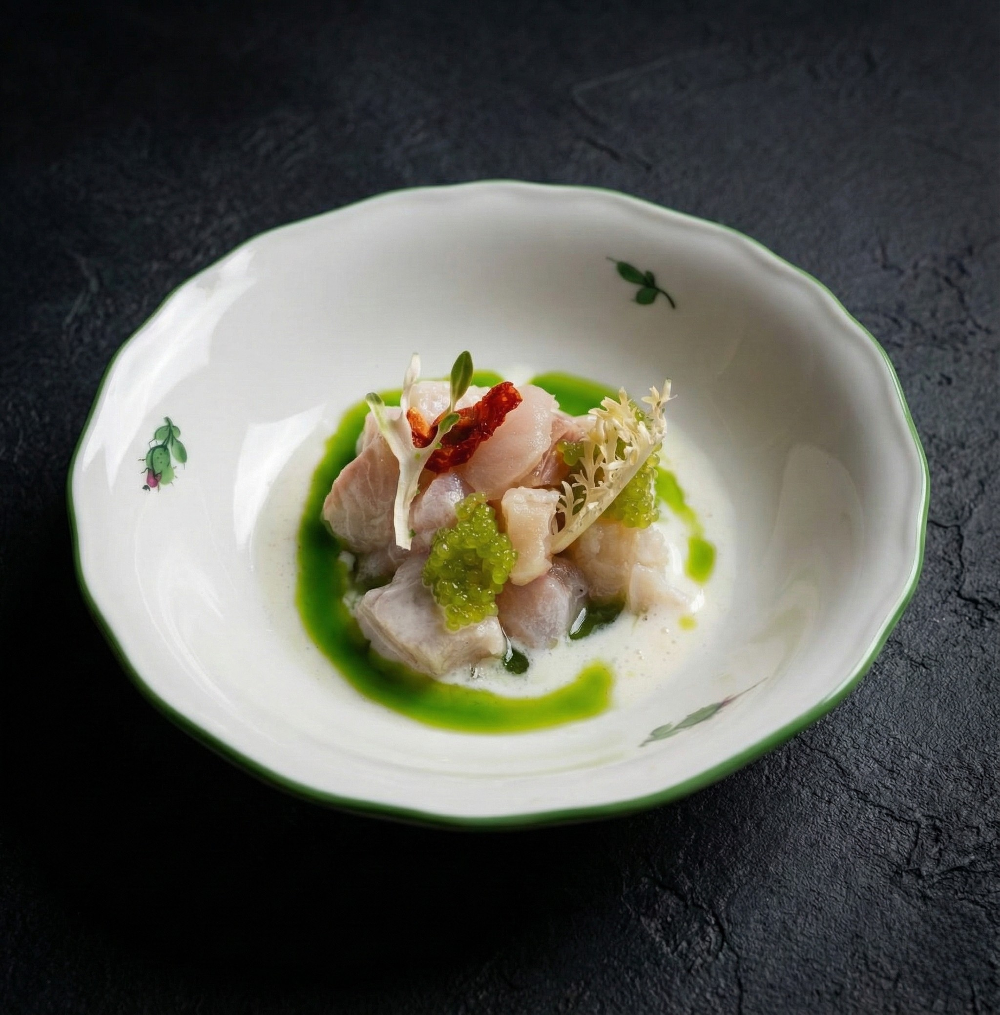 Gedämpfter Fisch mit grüner Soße, Garnelen, Meeresfrüchten und kleinen Kräutern in einer weißen Schüssel mit grüner Verzierung auf schwarzem Untergrund.