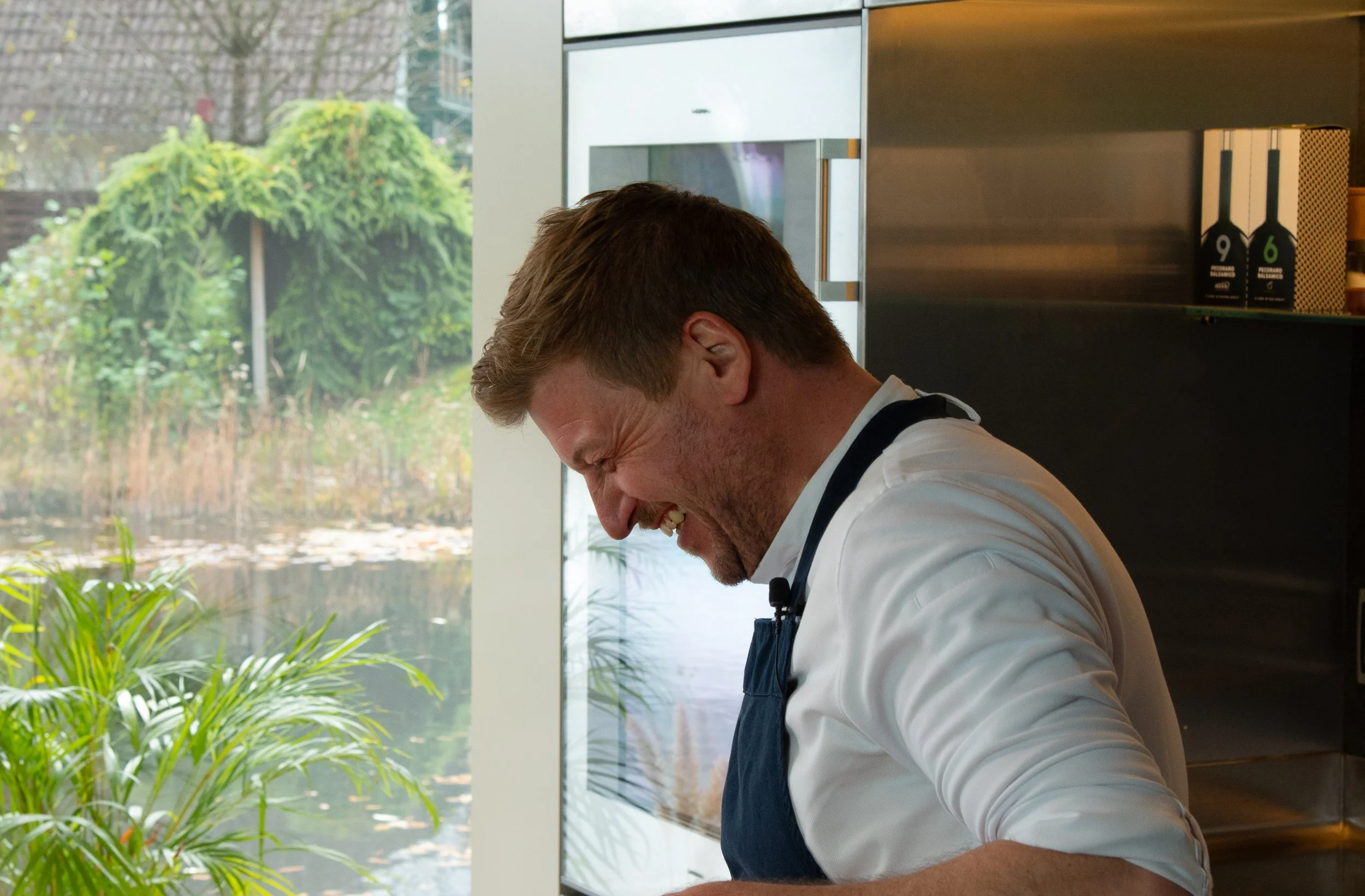 Ein Koch in einer weißen Jacke und dunkler Schürze, der vor einem Fenster mit Sicht auf einen Garten mit Büschen und Wasser steht, während er vor Lachen oder beim Kochen eine emotionale Reaktion zeigt.