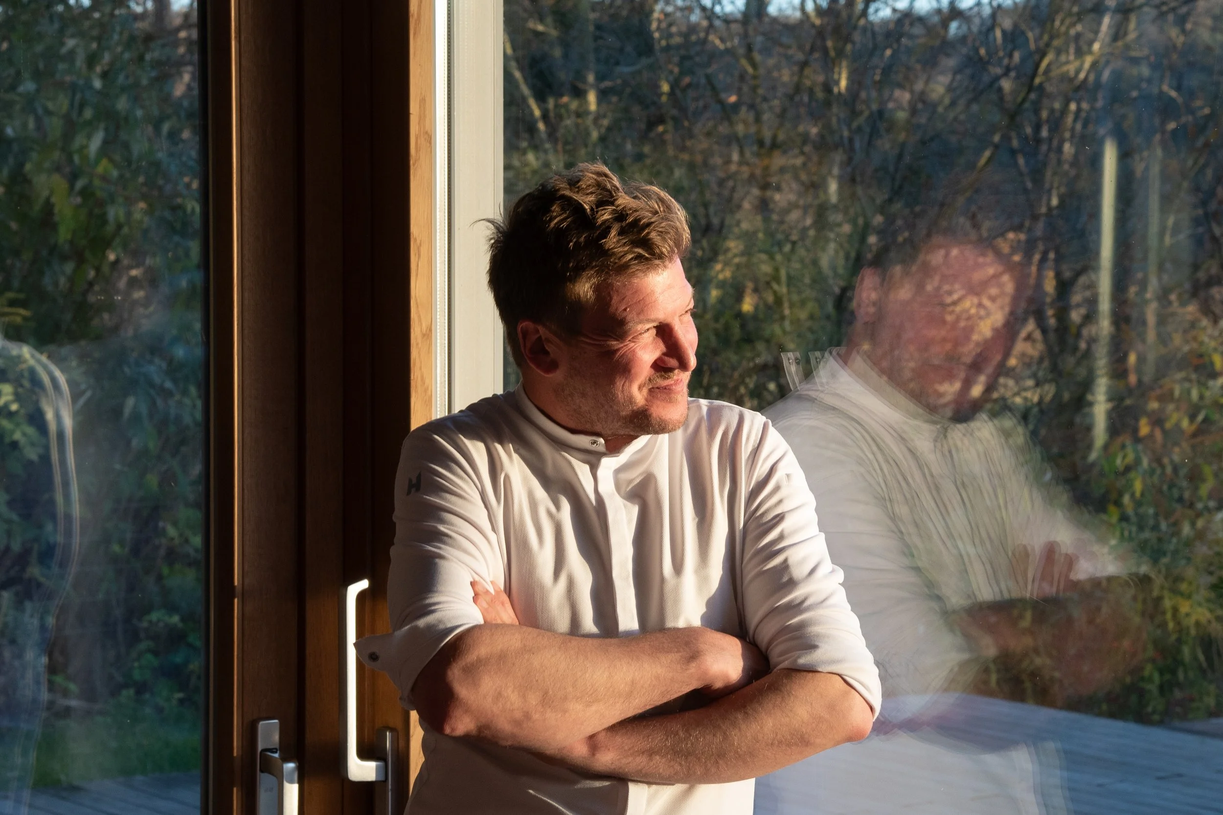 Mann schaut lachend durch ein Fenster mit Sonnenlicht, im Hintergrund Bäume im Herbst.