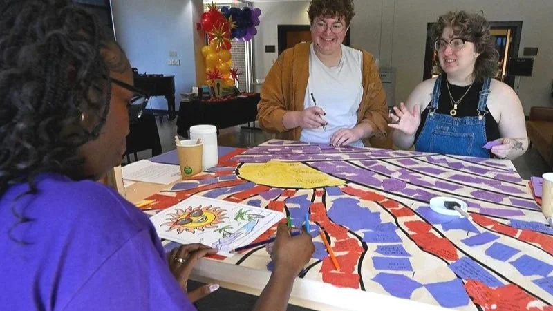 Three women creating and decorating a large colorful poster with balloons in the background.