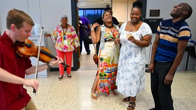 A group of people at an indoor airport or public space. A man is playing the violin, and three women and one man are smiling and laughing, enjoying the music and each other's company.