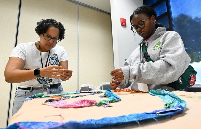 Two women standing at a table displaying colorful fabric with tied designs, working on a craft project.
