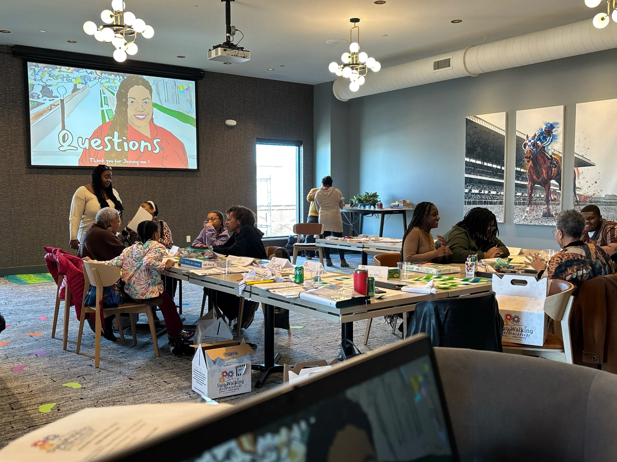 People sitting at tables in a conference room, engaging in discussion. A woman standing near a table, a large screen displaying an illustration of a woman with the word 'Questions', and two large artwork panels of a horse race on the wall.