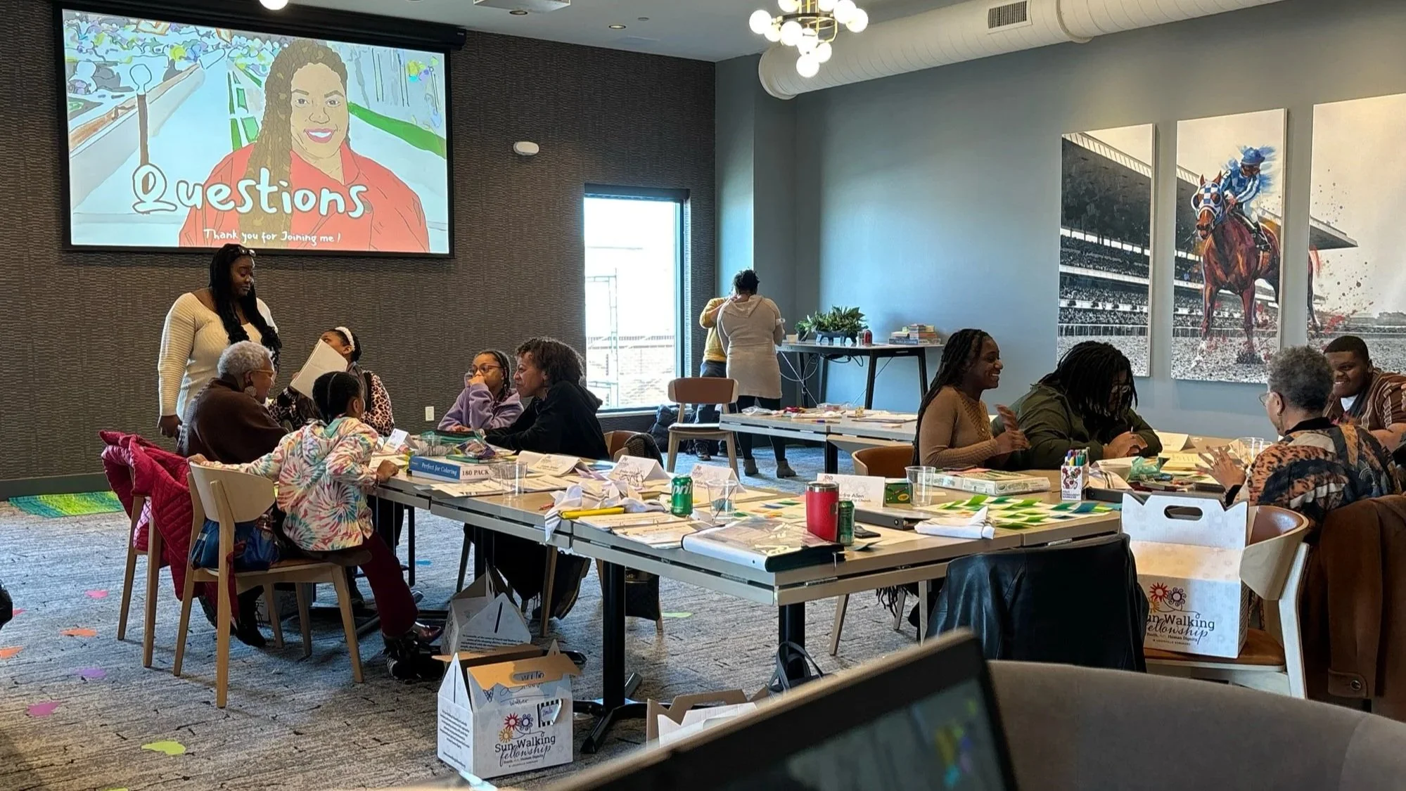 A group of people sitting around tables in a conference room, engaging in a workshop or meeting. There is a large screen displaying an illustration of a woman with the word 'Questions' and the phrase 'Thank you for joining me!' in the background. Some participants are talking and interacting, with various materials and drinks on the tables.