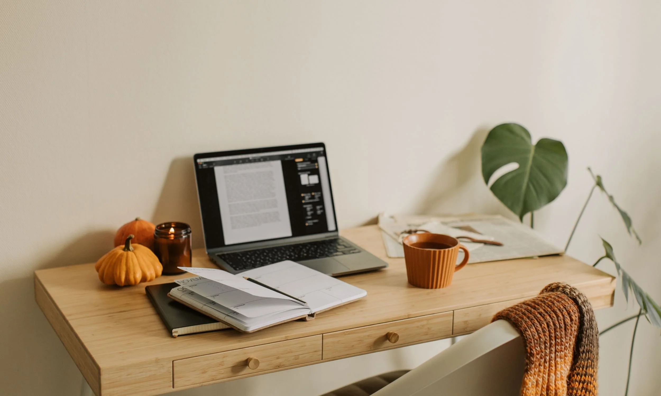 A cozy wooden desk with a laptop, a spiral planner, a brown and orange striped mug, small pumpkins, a candle, and a brown knitted blanket with a leafy plant in the background.
