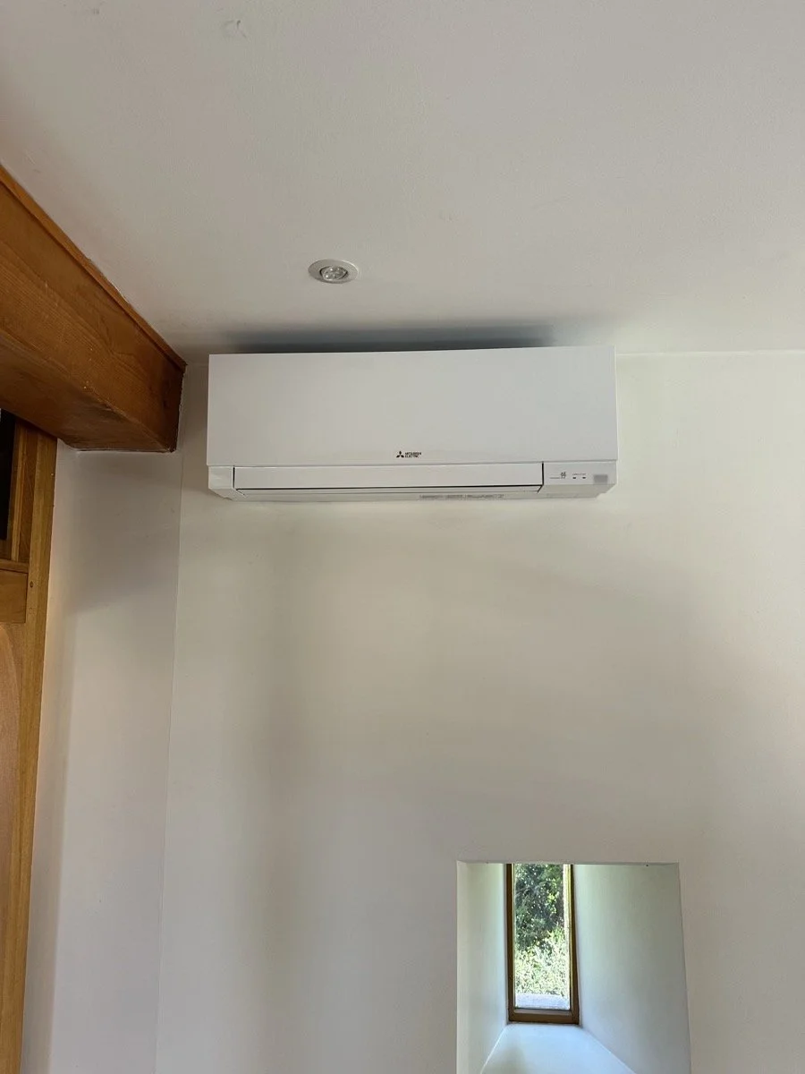 White wall-mounted air conditioner unit in a room with a white ceiling, a small window showing trees outside, and a wooden beam on the left side.