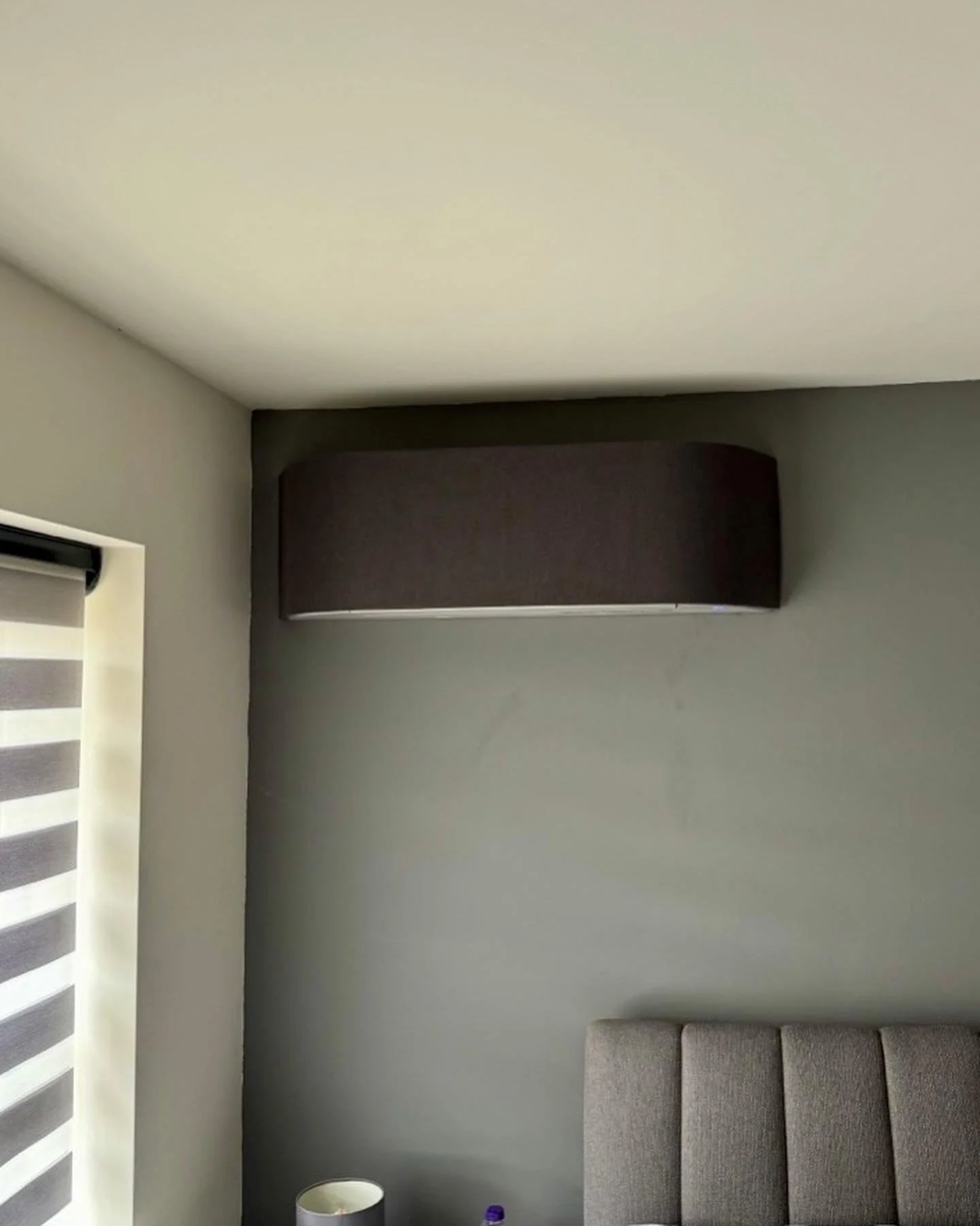 Indoor room corner with gray wall, beige ceiling, gray upholstered bed headboard, window with black and white striped blind, and black wall-mounted air conditioning unit.