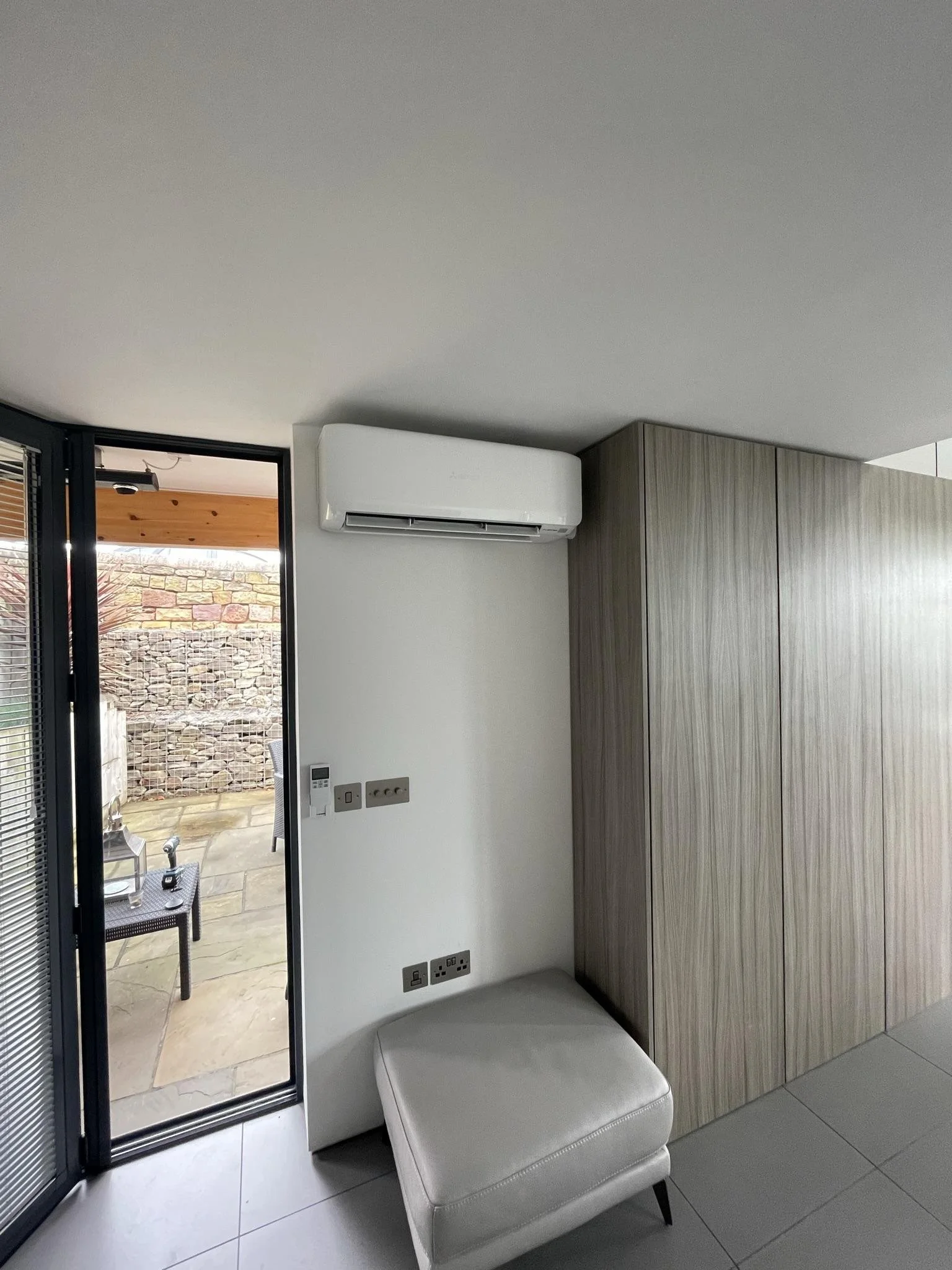Interior view with a white air conditioning unit mounted on a white wall, beside a wooden cabinet, and a small white ottoman below. There is a glass door leading to an outdoor patio with a stone wall.