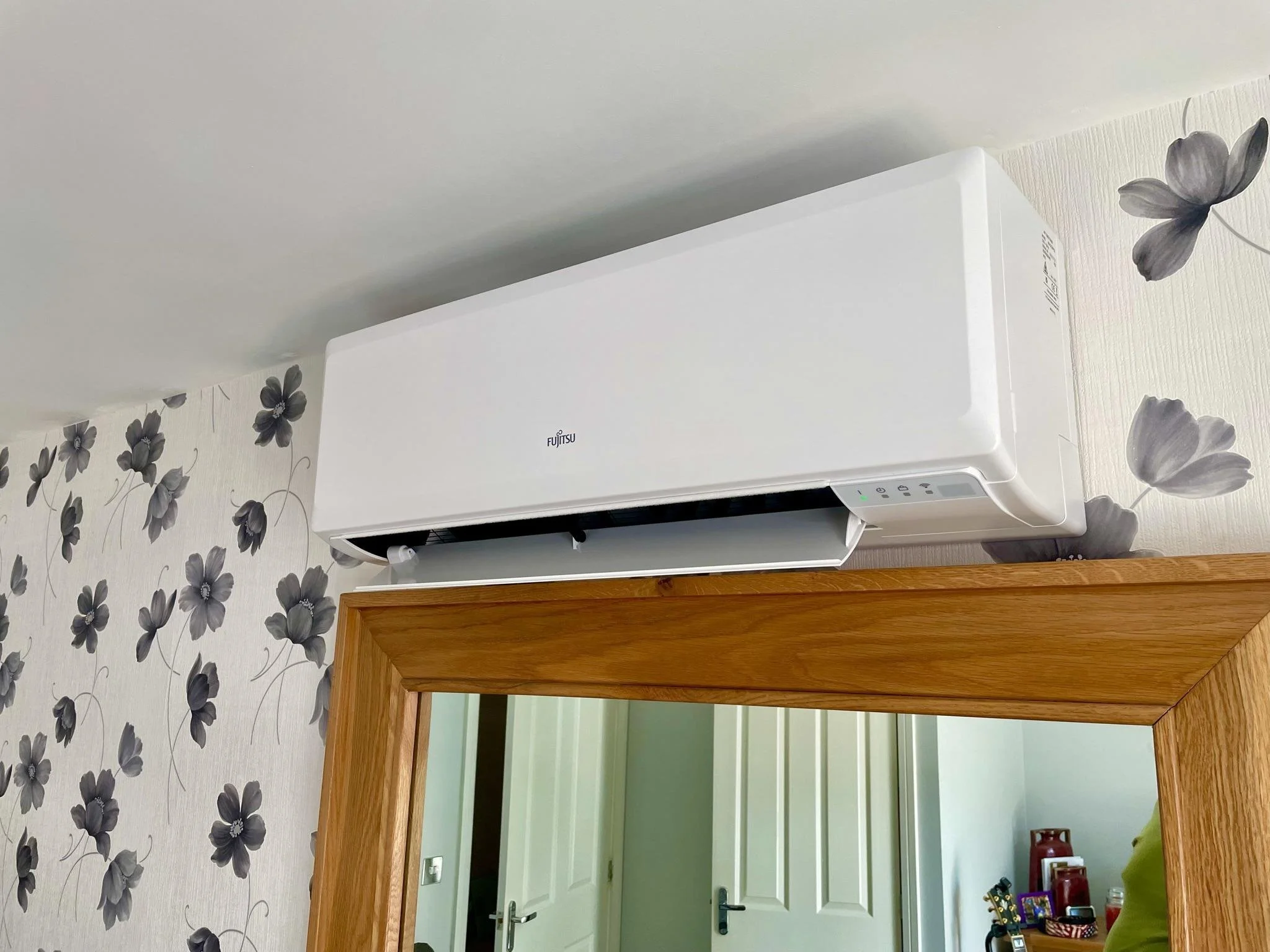 White Fujitsu wall-mounted air conditioner installed above wooden doorway, with floral patterned wallpaper and partially visible room with door and various objects inside.