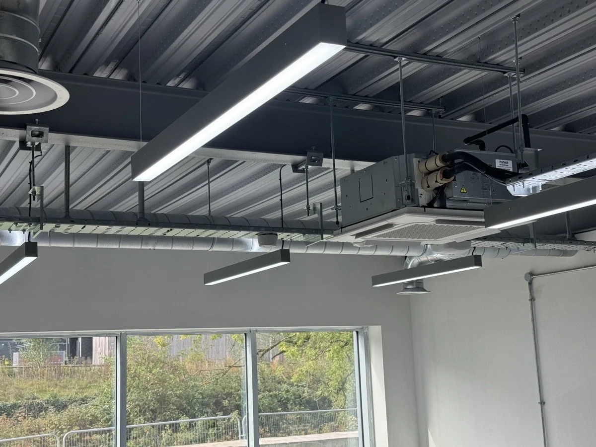 Ceiling with suspended industrial lighting, HVAC ductwork, and electrical systems in a modern office with large windows and outdoor view.