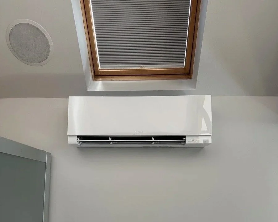 White wall-mounted air conditioner unit below a skylight window with closed blinds, and a round ceiling vent in the ceiling.