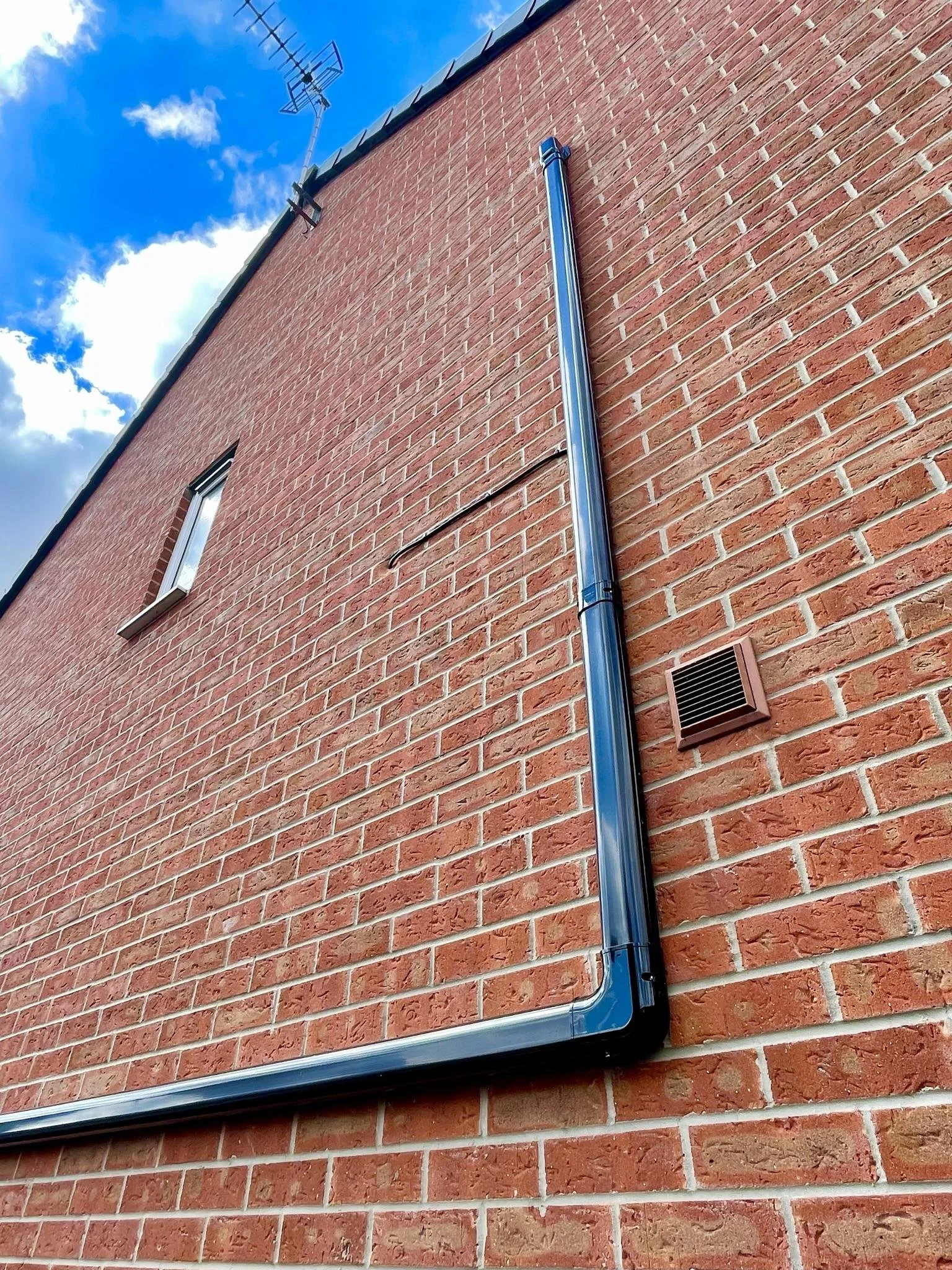 Close-up of a brick building exterior with a shiny metal pipe running vertically along the wall, a small window, a ventilation grate, and antenna on the roof under a partly cloudy sky.