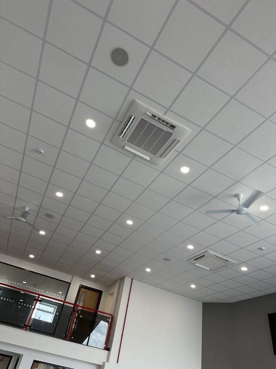 Ceiling with fluorescent lights, ceiling fans, an air conditioning vent, and acoustic ceiling tiles in a modern building interior.